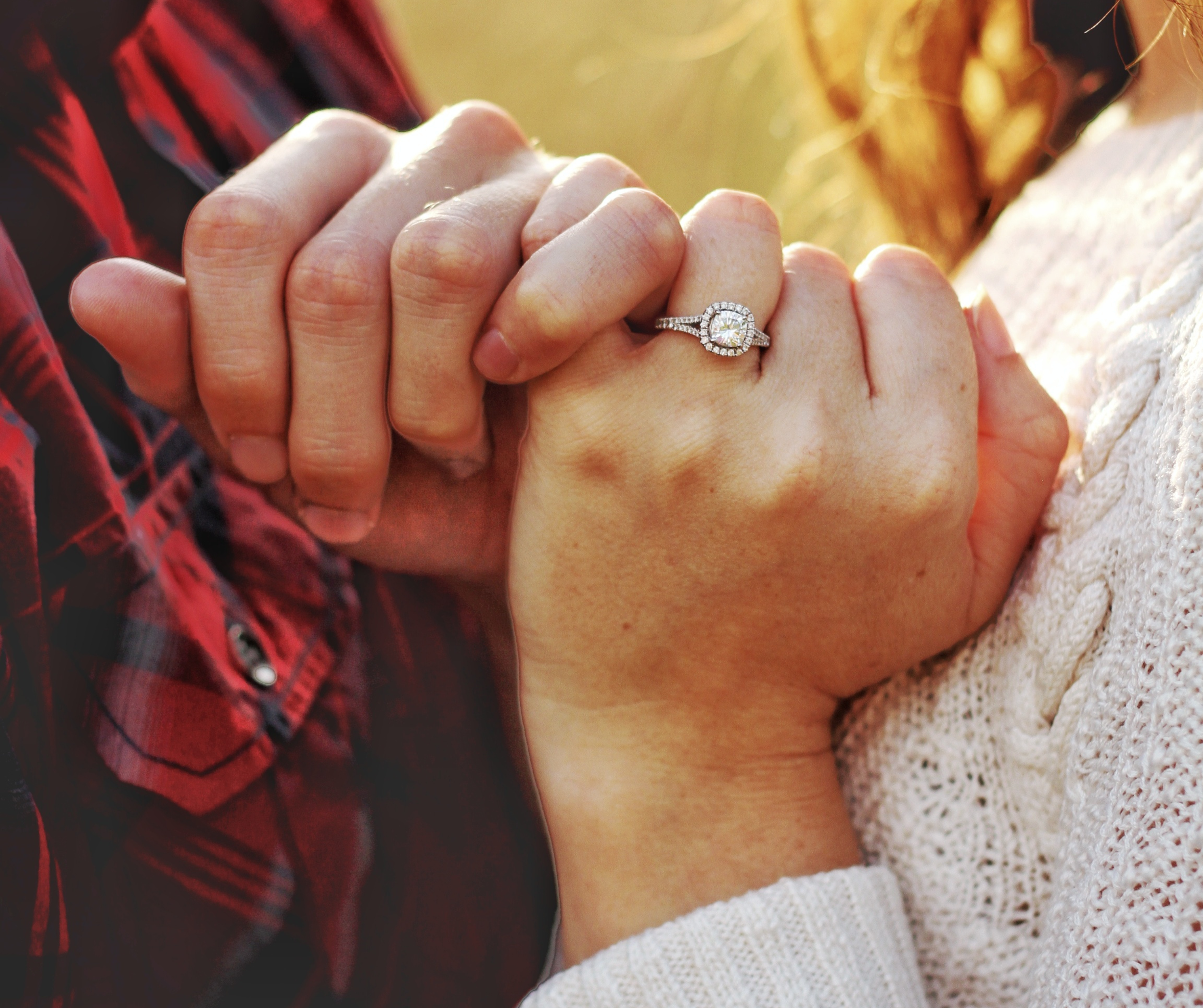 Close-up of two hands clasped together, with a diamond engagement ring on the woman's ring finger, set against a blurred outdoor background.