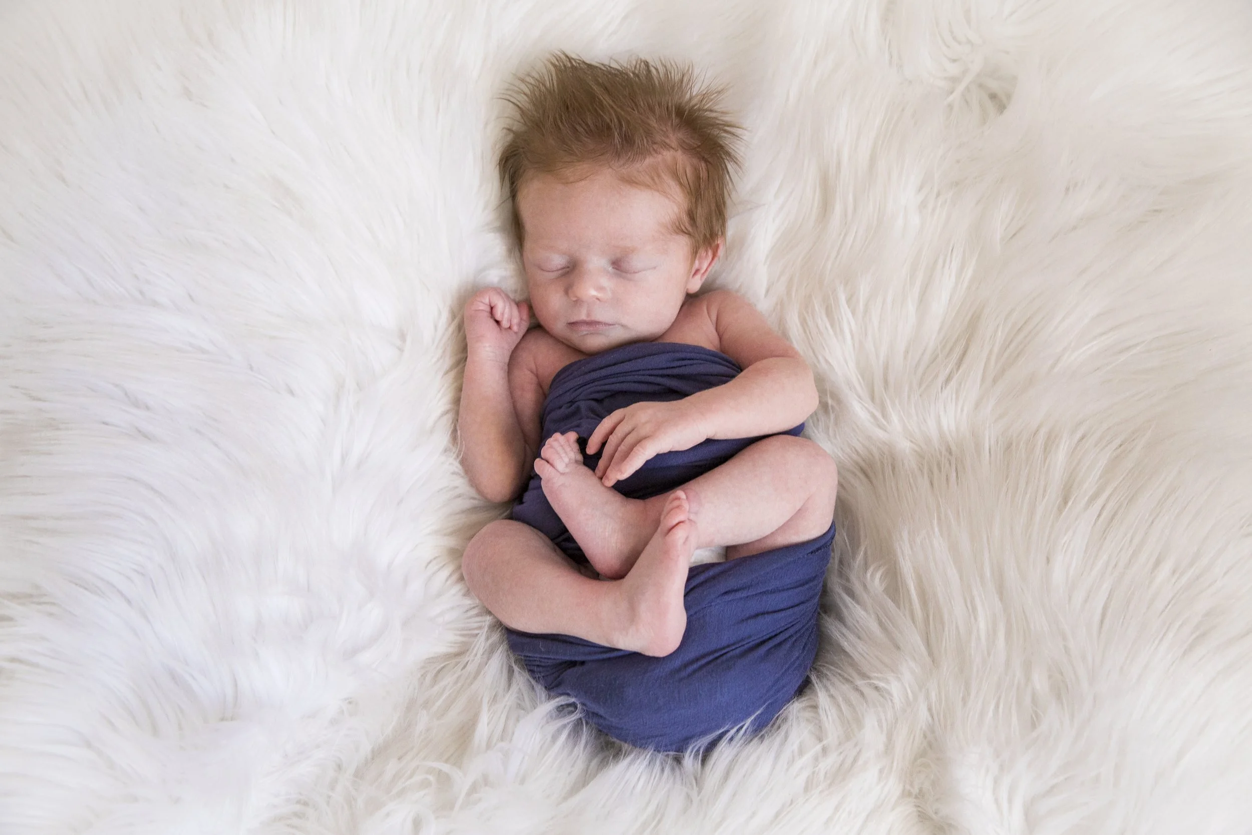 Newborn baby with red hair sleeping on a white furry blanket, wrapped in a navy blue cloth.
