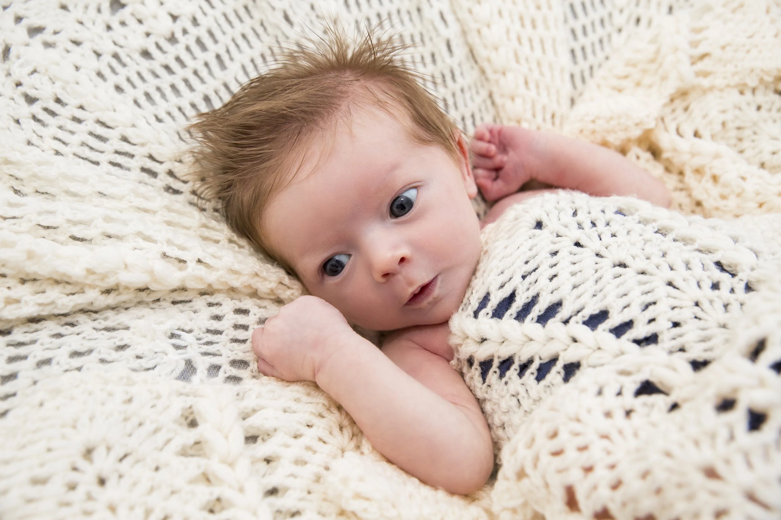 A baby with light skin and blue eyes lying on a cream-colored knitted blanket, looking towards the camera.
