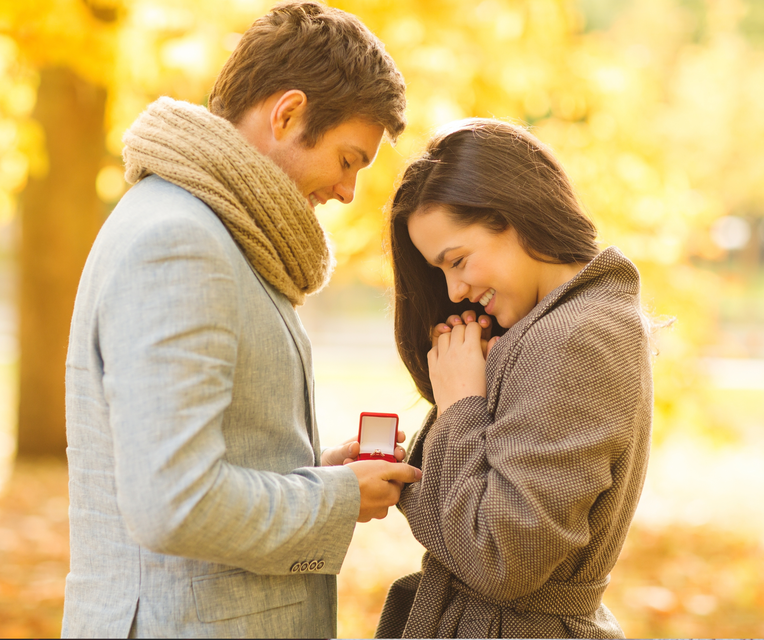 A man on one knee proposing to a woman in a park with autumn leaves, with the woman smiling happily and clasping her hands together.