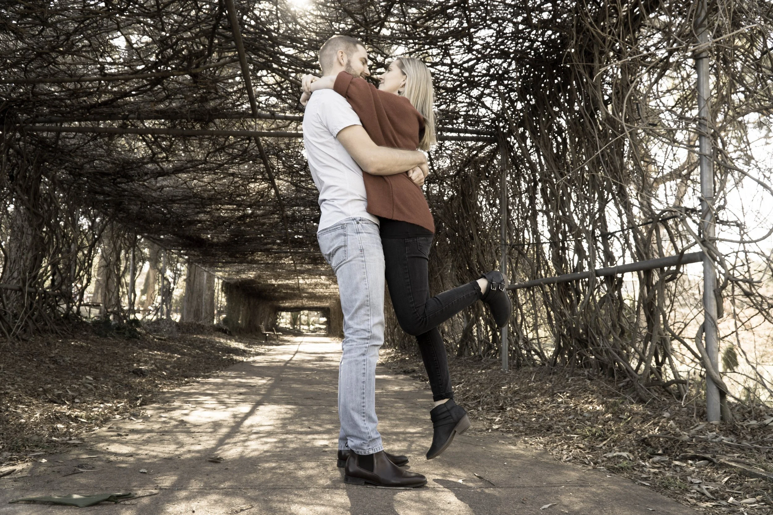 A couple embracing and kissing on a dirt path under a tunnel of intertwined trees and branches.