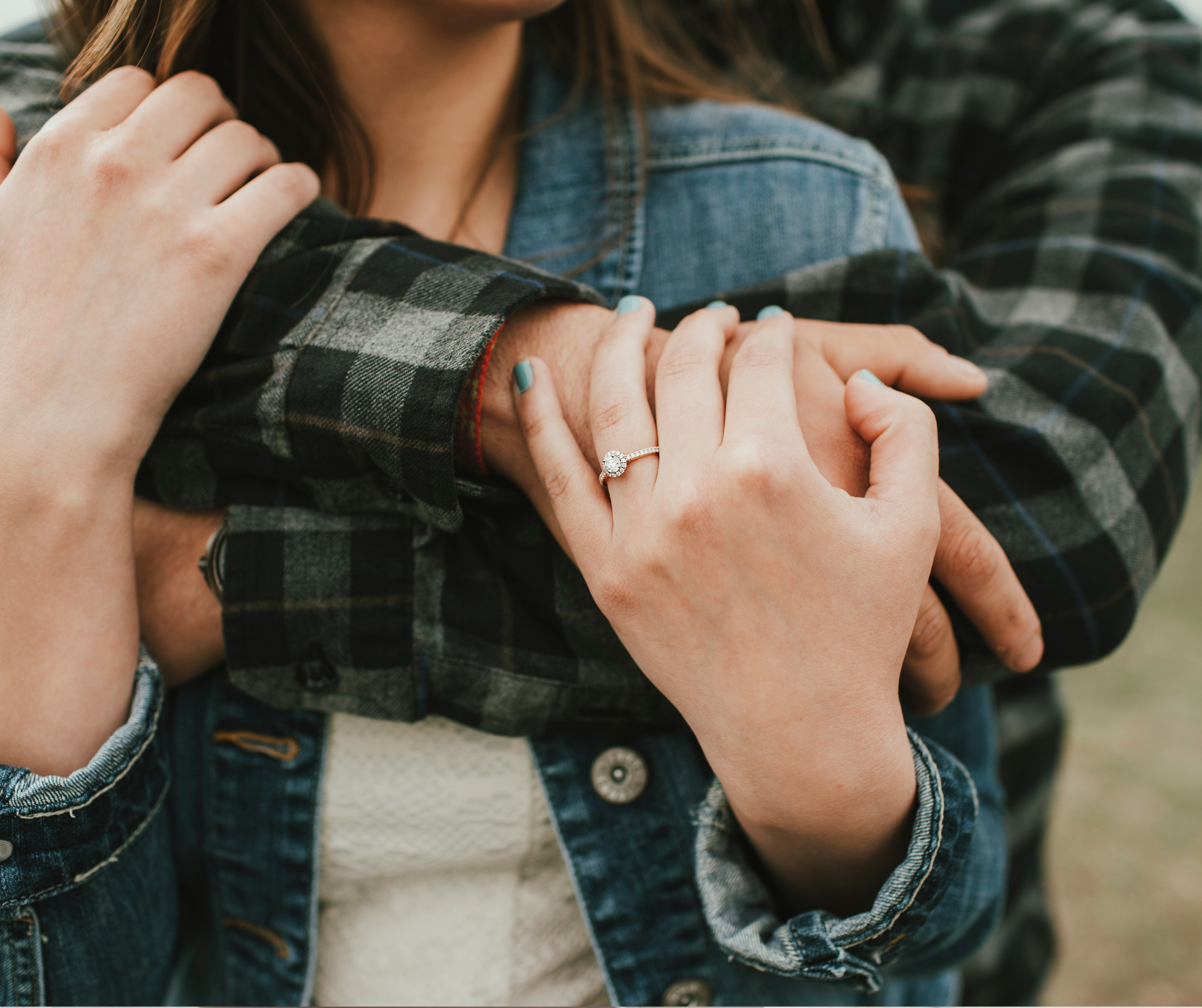 Close-up of a woman's left hand showing an engagement ring, as she embraces a person wearing a green and black plaid shirt with her right hand. The background is blurred, focusing on the hands and ring.