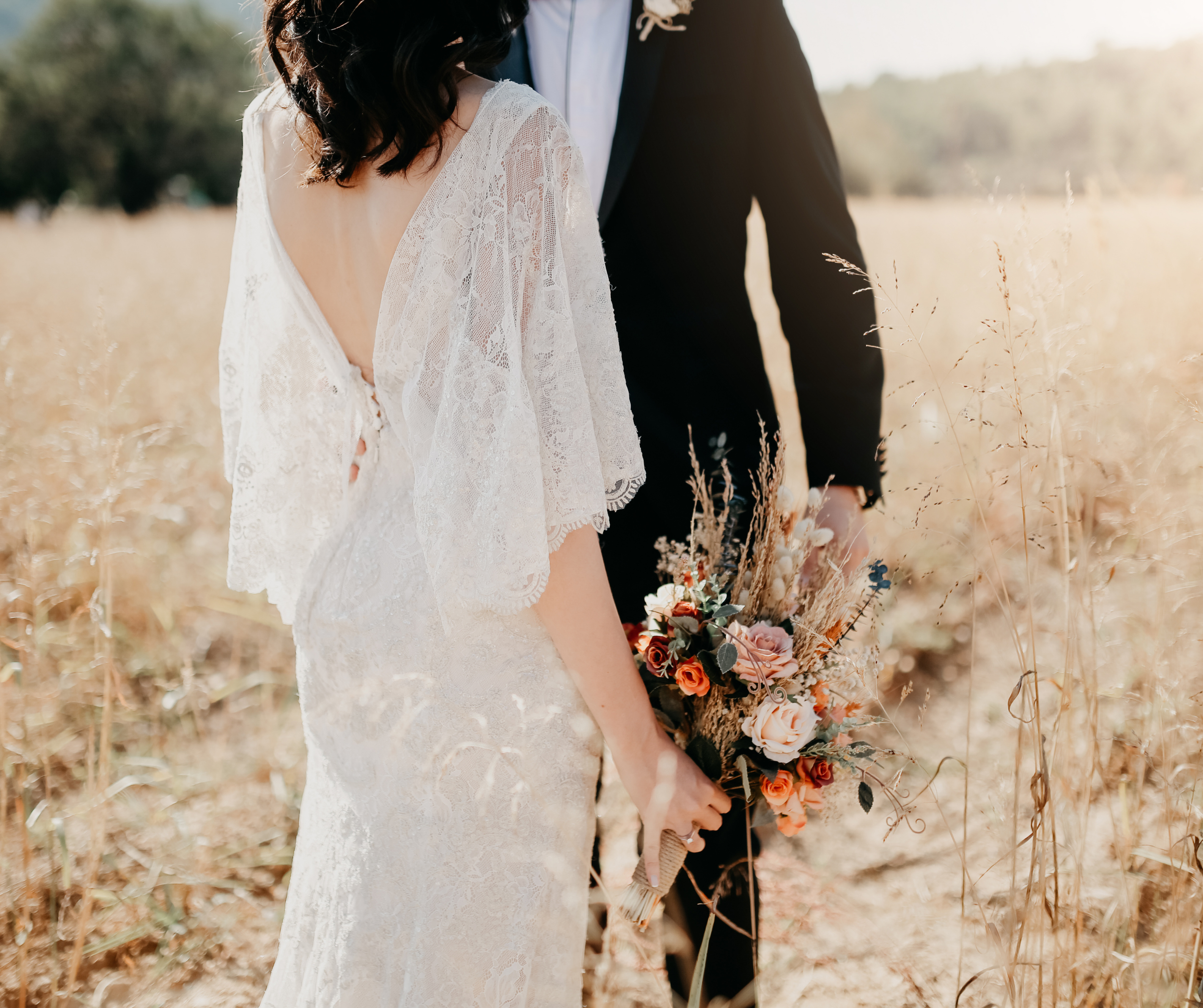 Bride in lace wedding dress holding bouquet in field, groom in suit standing beside her.