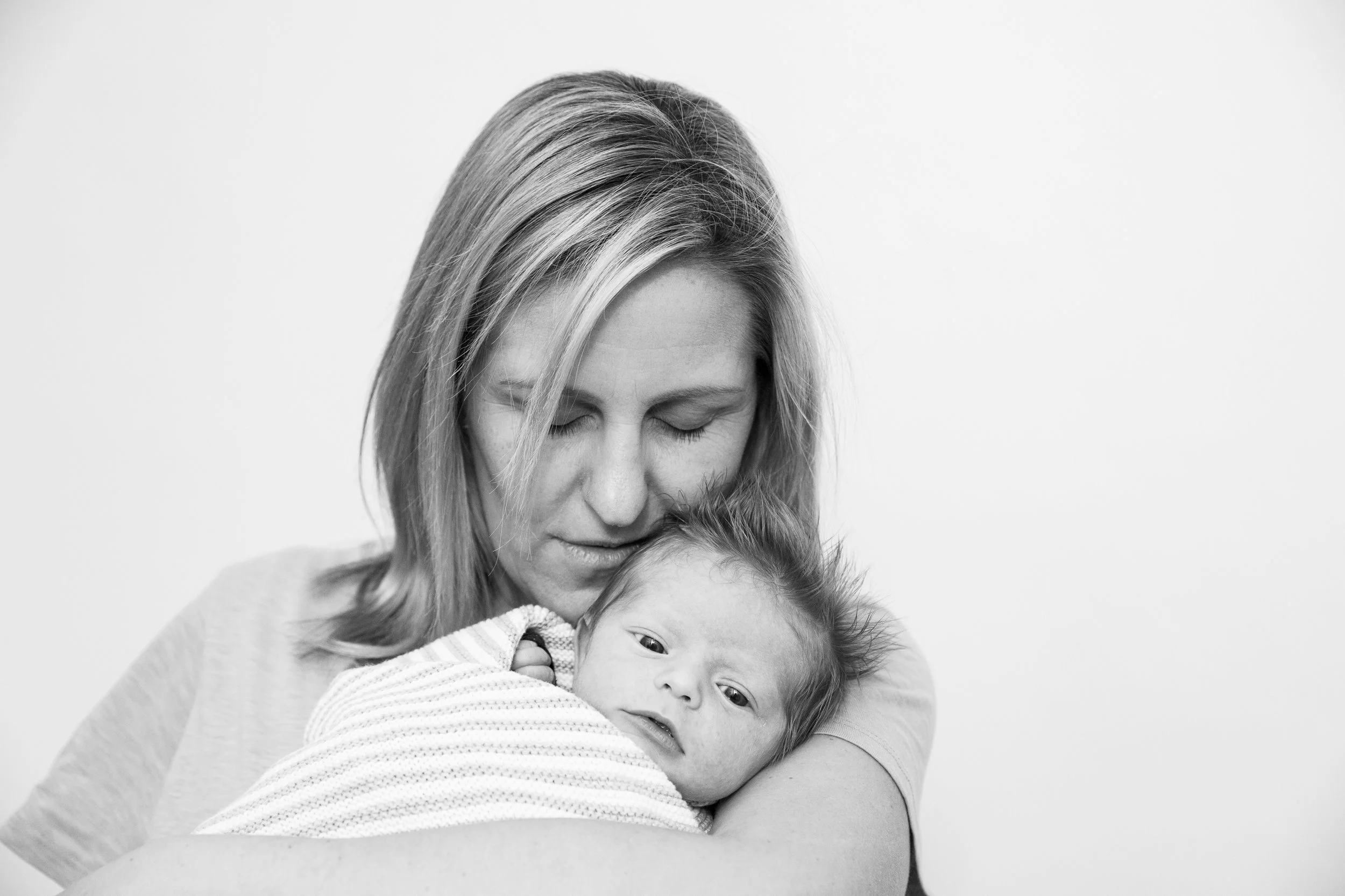 Black and white photo of a woman hugging a young child, with her eyes closed and head resting on the child's head, against a plain background.