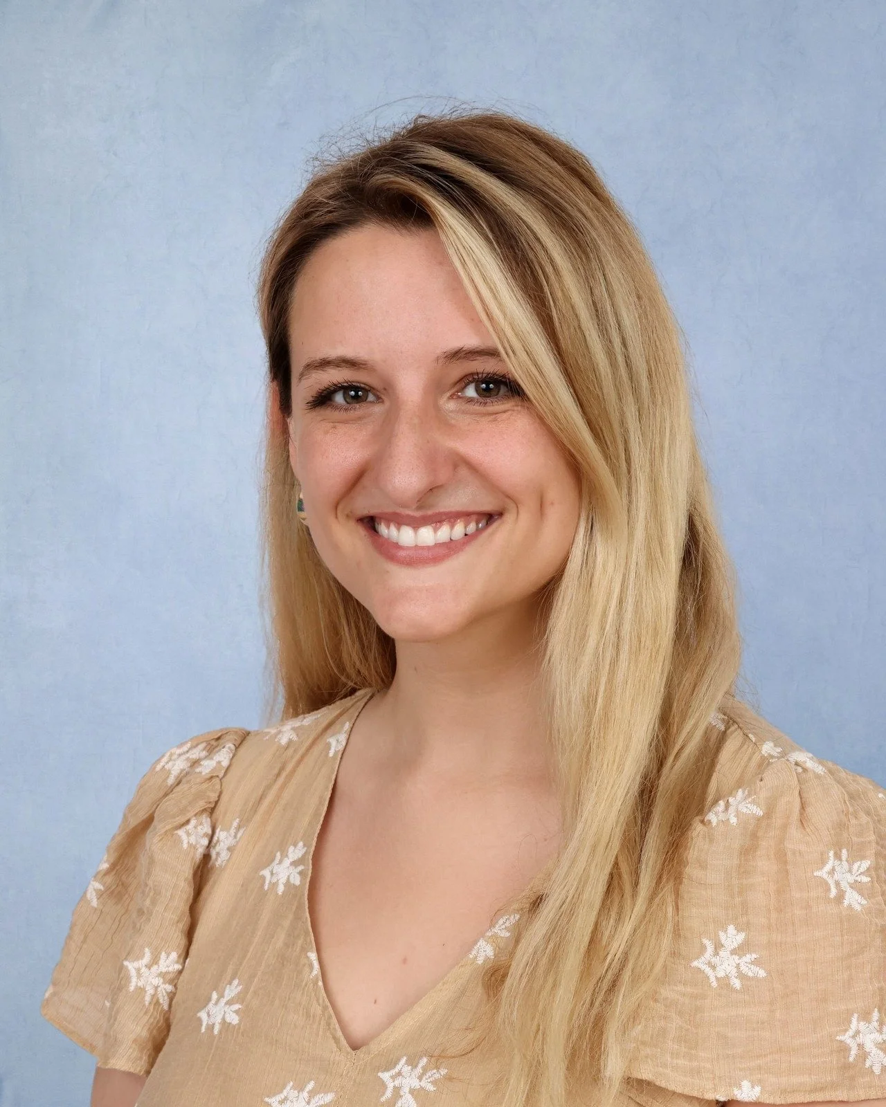 A smiling young woman with shoulder-length blonde hair wearing a pink sweatshirt with white text, standing against a plain light blue background.
