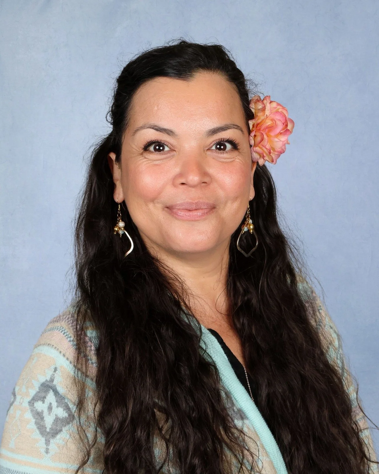 A woman with long, curly dark hair, smiling, wearing a turquoise floral blouse and white earrings, against a light blue background.