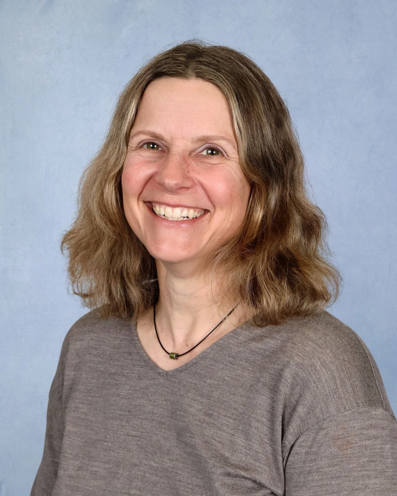 A woman with long dark hair, wearing a brown top, standing in front of a light blue background.