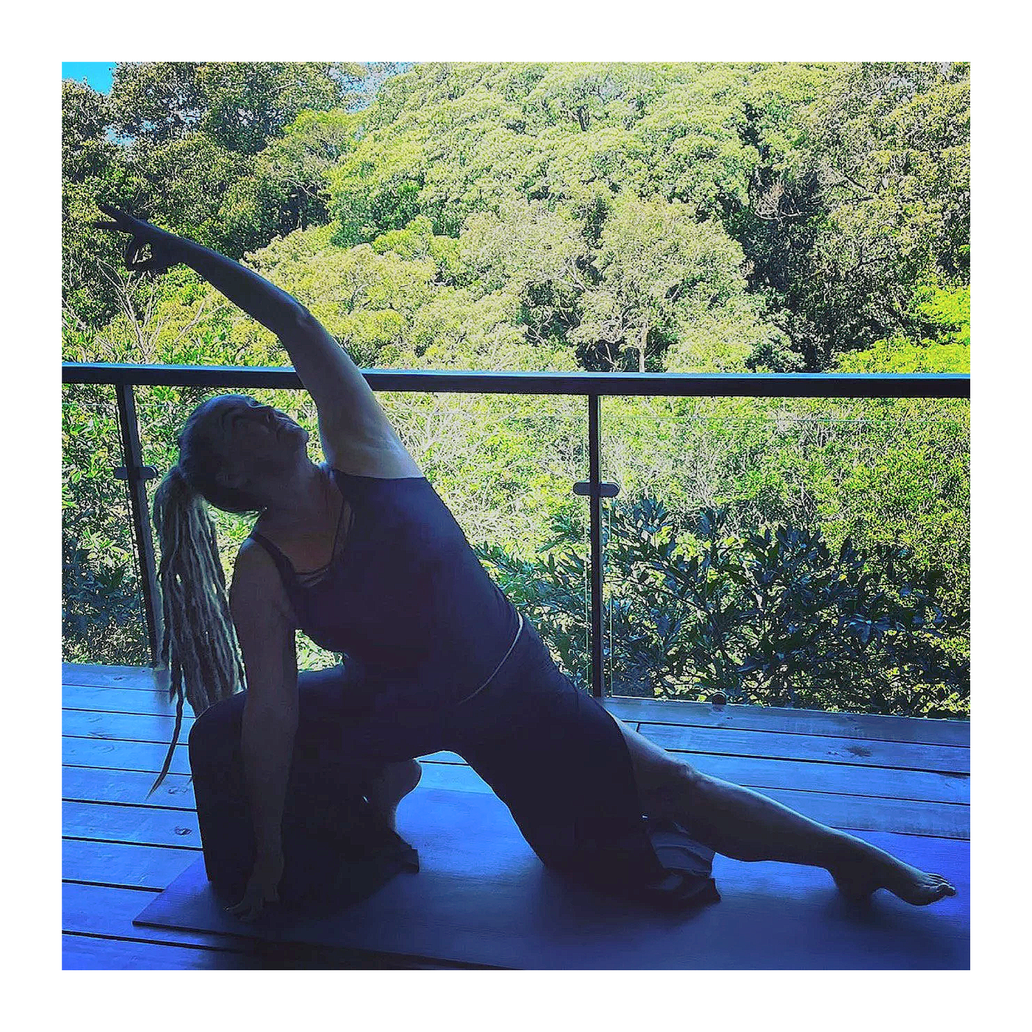 A woman with dreadlocks sitting cross-legged on a purple yoga mat, practicing meditation on a colorful, flower-patterned floor. She's surrounded by singing bowls, and has a large floral tattoo on her back.