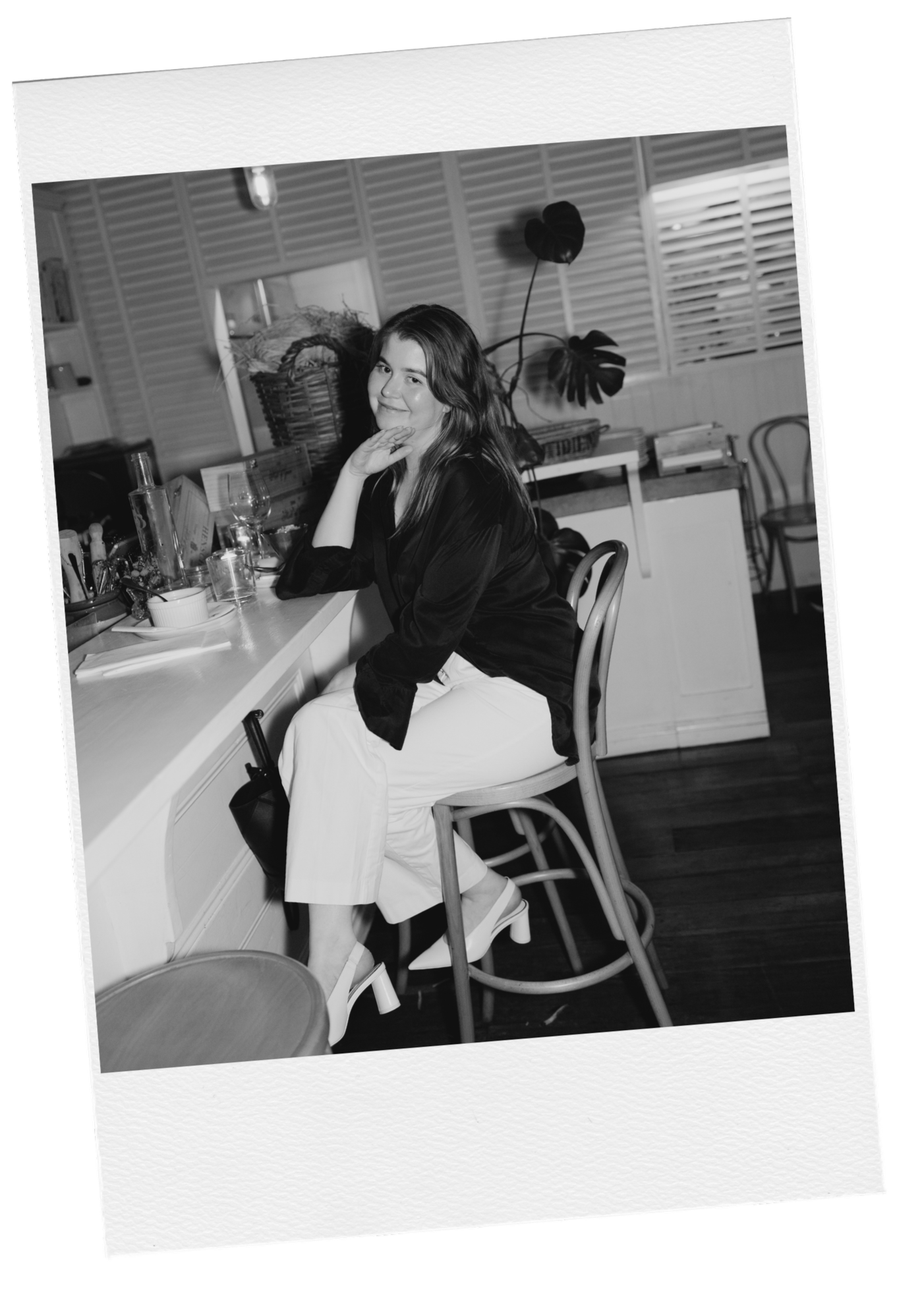 A woman sitting on a barstool at a counter in a cafe or restaurant, smiling. She is wearing a dark top and white pants. The counter has glasses, a bottle, and other items. The background has a large leafy plant and window shutters.