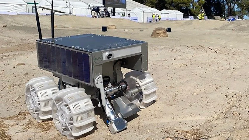 A small robotic vehicle with six large white wheels, a box-like top housing solar panels, and various mechanical components, on a sandy outdoor area with tents in the background.