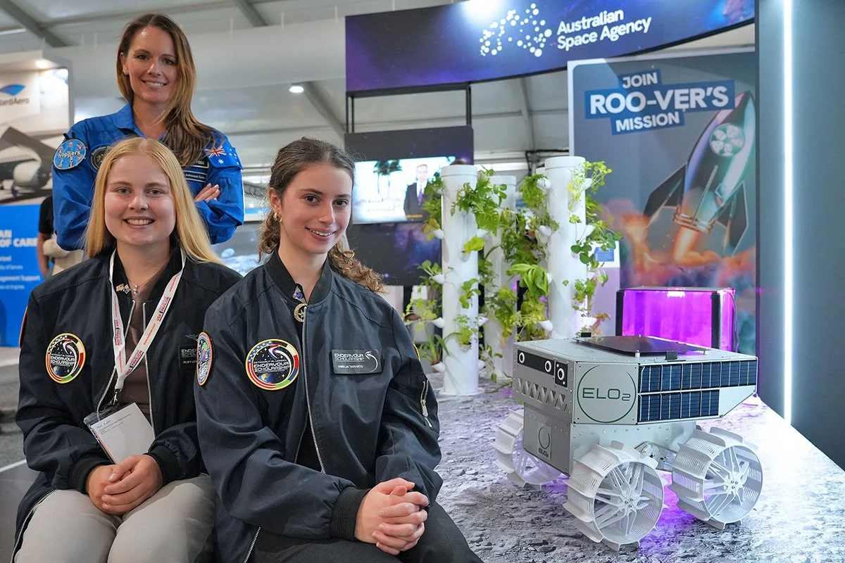 Three women in NASA and space agency uniforms posing at a space exhibit. One woman stands behind, and two women sit in front, with a robotic rover model labeled 'ELO2' and plants in the background.