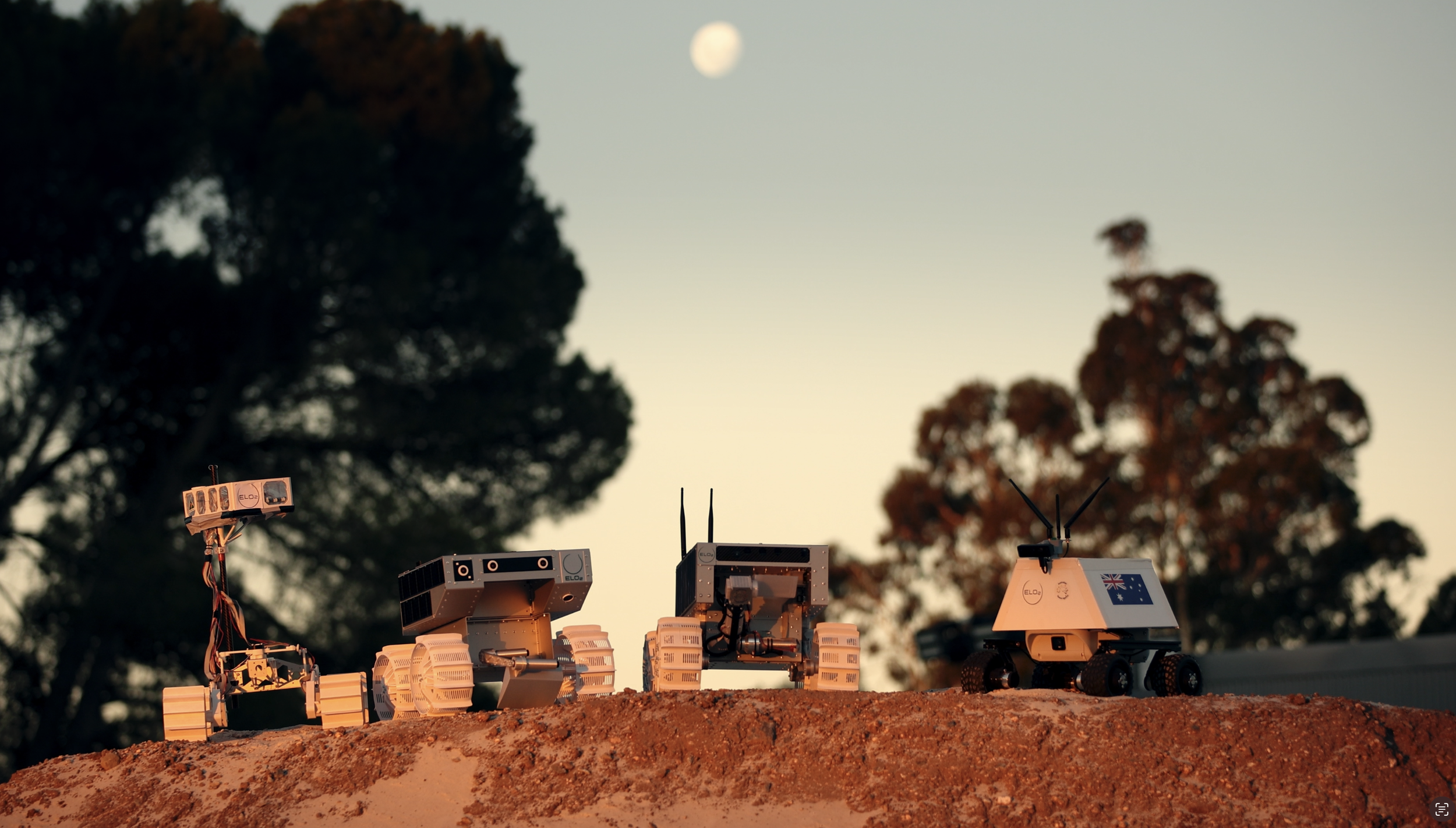 A small robotic rover with white wheels and a rectangular body on a dirt surface outdoors, with tents and people in the background.