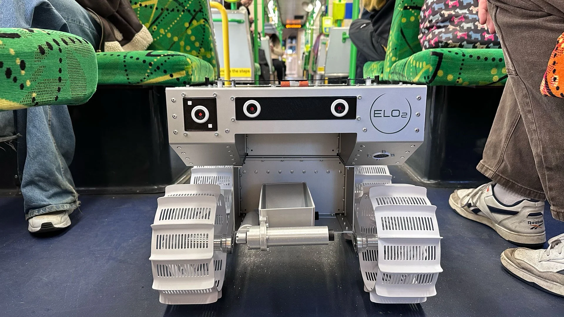 A robot with caterpillar tracks is located inside a bus, between seats with students sitting on either side. The robot is labeled 'ELO2' and has a box-like structure with sensors and controls on top.