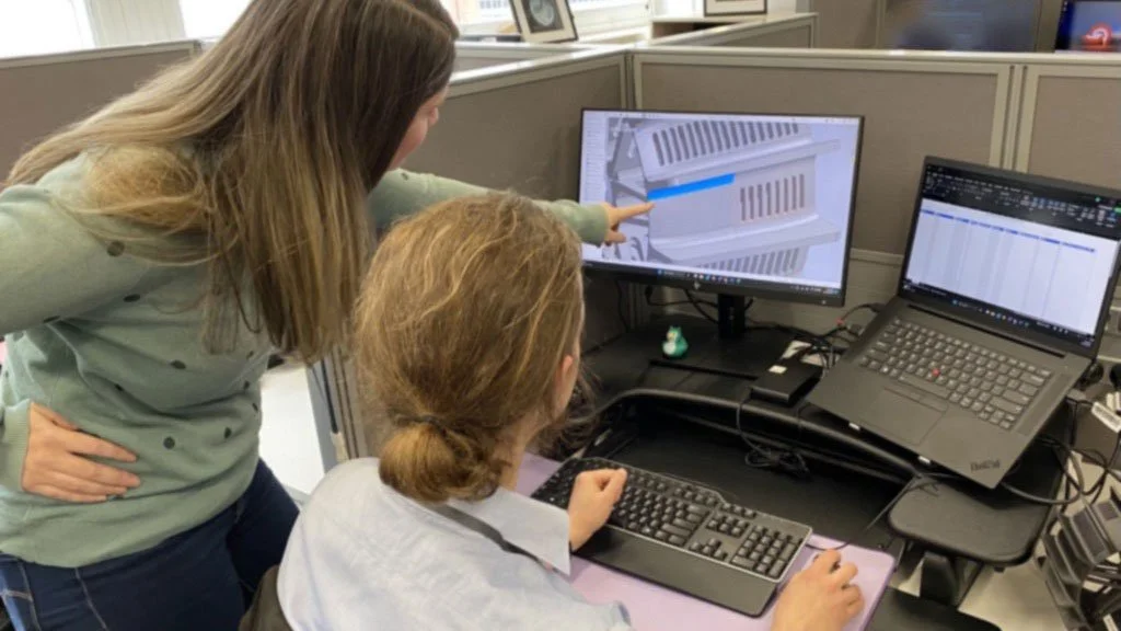 Two women working at a computer with technical drawings and graphs on the monitors in an office environment.
