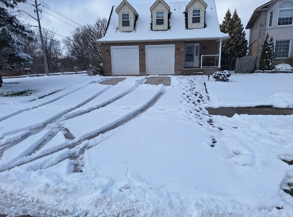 Snow-covered driveway leading to a house with a garage, tire tracks and footprints in the snow.