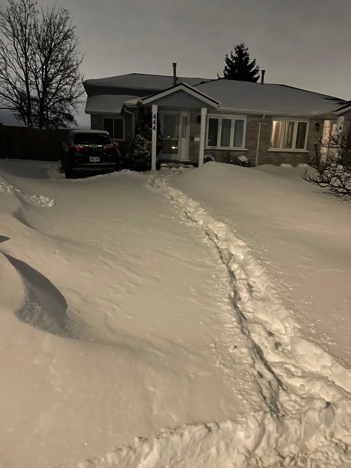 A snow-covered driveway leading to a house with a car parked in front. Footprints and tire tracks are visible in the snow, with a house and trees in the background during dusk or dawn.