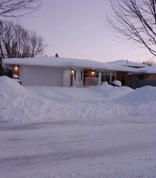 A house surrounded by snow with snow-covered driveway and trees in winter, porch lights on, during dusk or early evening.