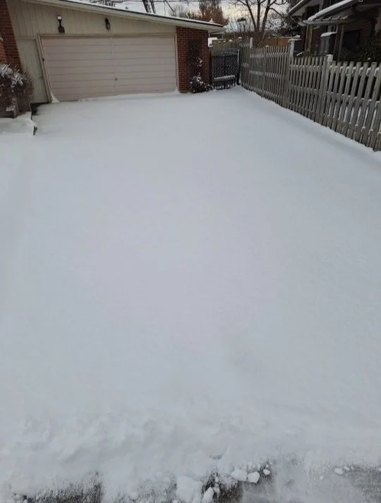 Snow-covered backyard with a garage, wooden fence, and snow on the ground and driveway.
