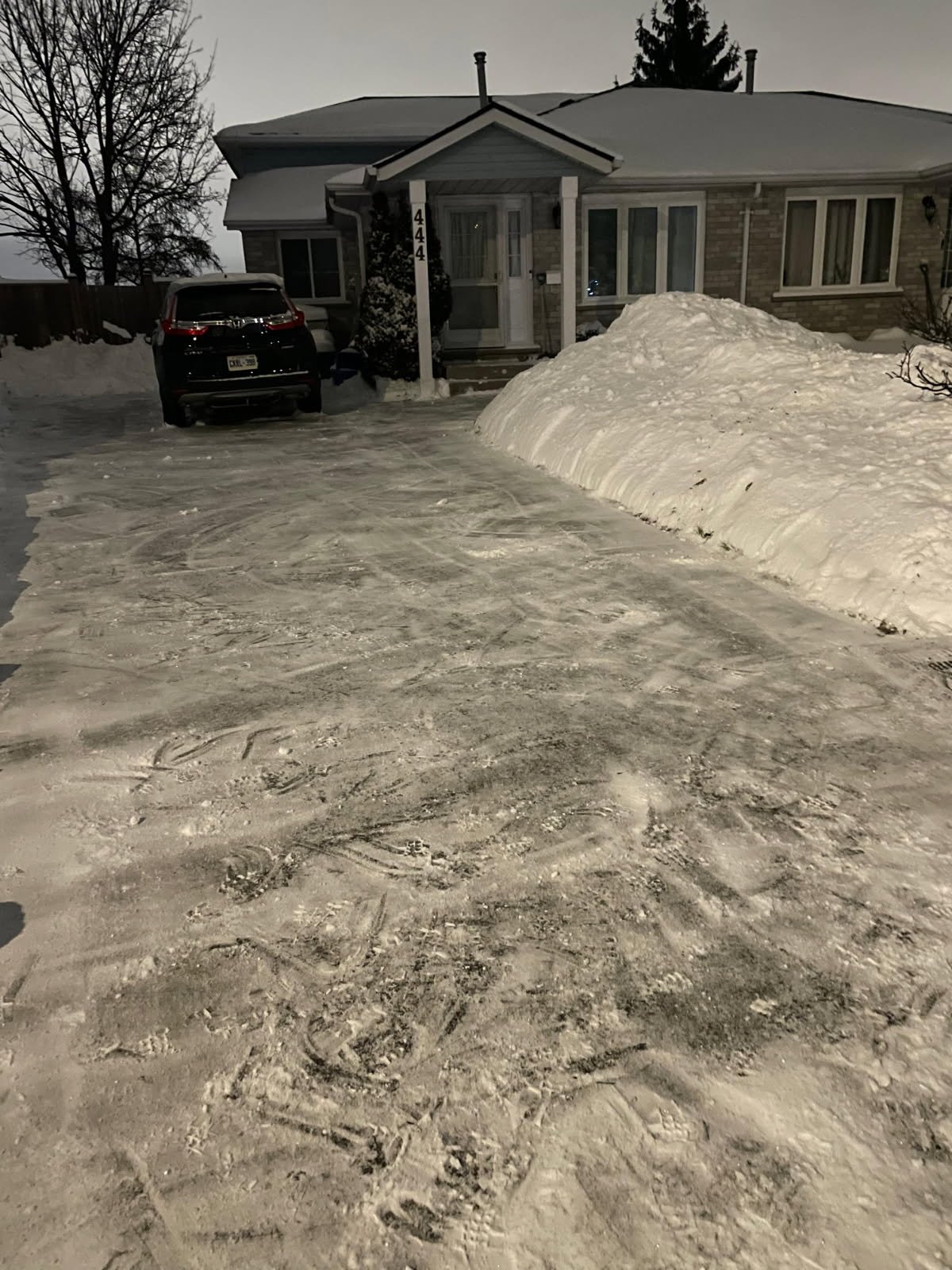 A snow-covered driveway leading to a house with a black SUV parked in front. There is a large snowbank on the right side of the driveway and tire tracks on the snow.
