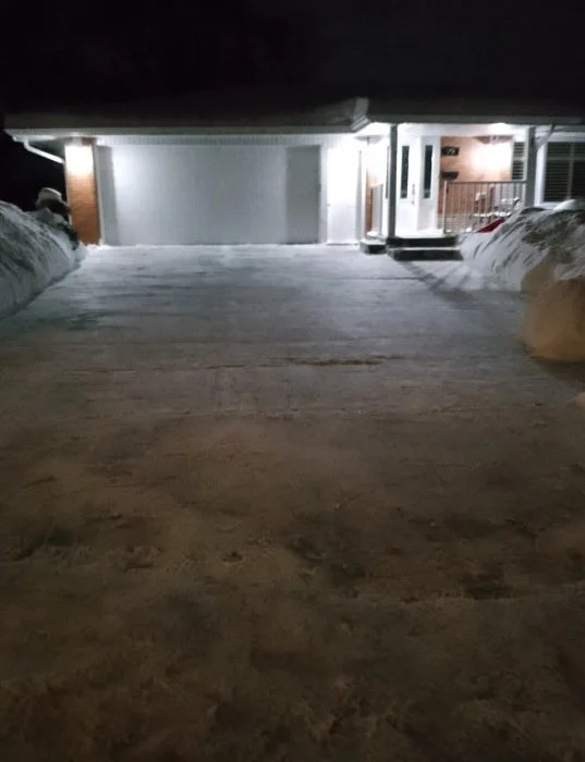 Snow-covered driveway leading to a well-lit house at night.