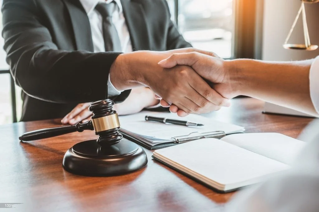 Two people shaking hands across a legal or business desk with a gavel, papers, and a pen on the table.