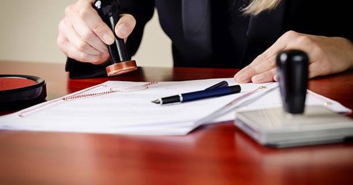 Person stamping documents on a wooden desk with a black ink pad, pen, and notepad nearby.