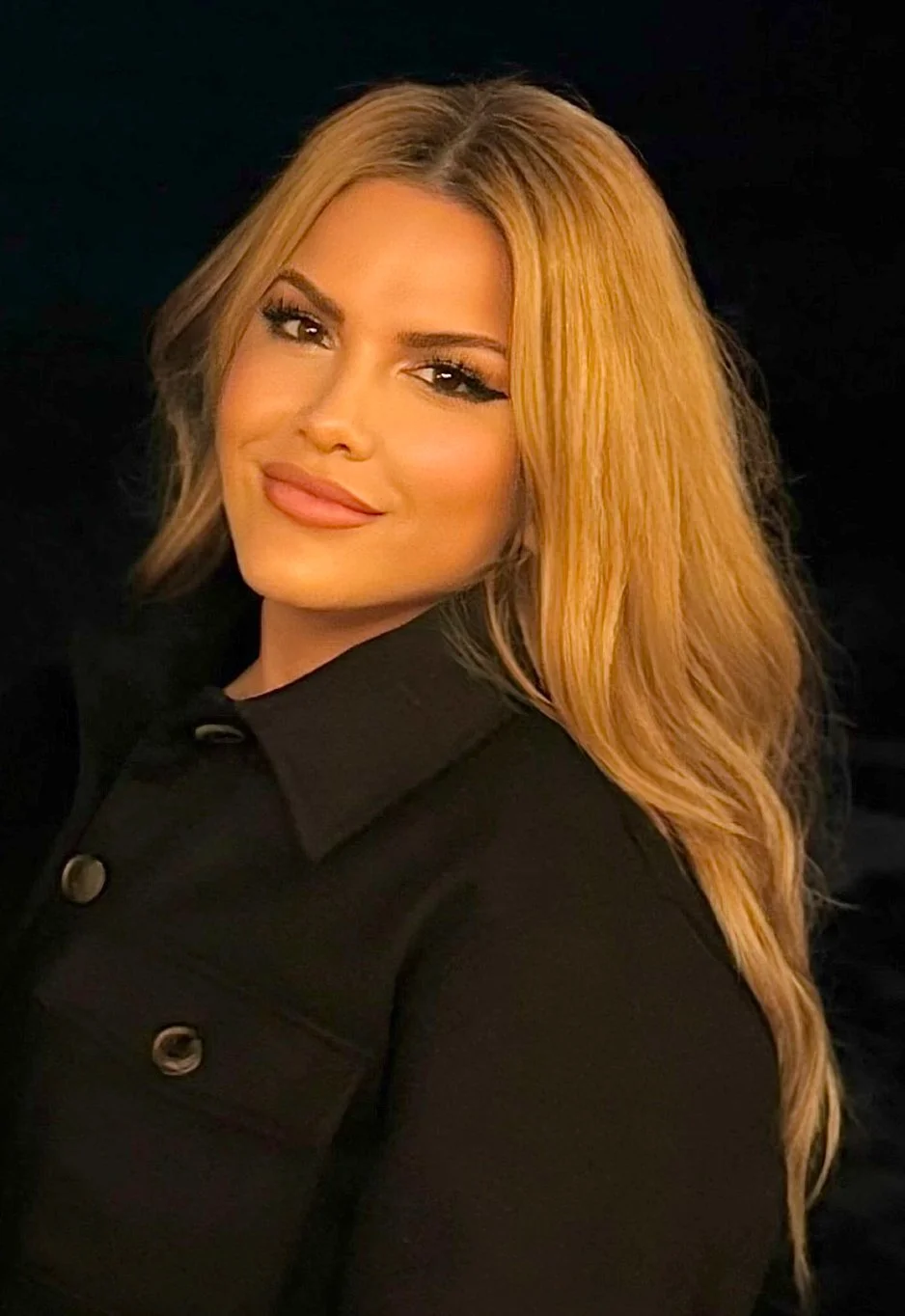 A woman with long wavy blond hair, wearing makeup, and a black button-up shirt, looking at the camera with a slight smile against a dark background.