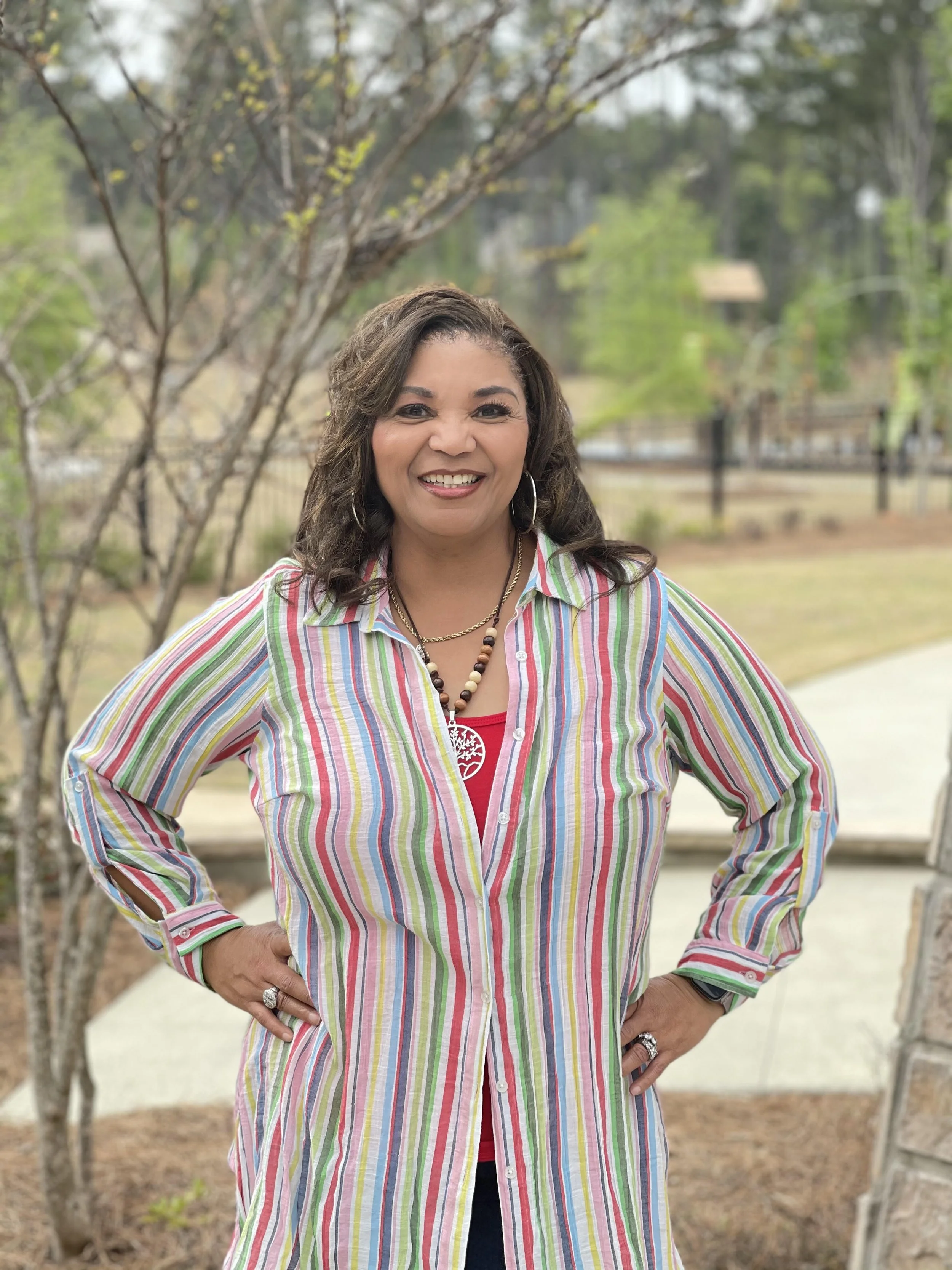 A smiling woman standing outdoors with trees and a fence in the background, wearing a colorful striped shirt, jewelry, and her hands on her hips.