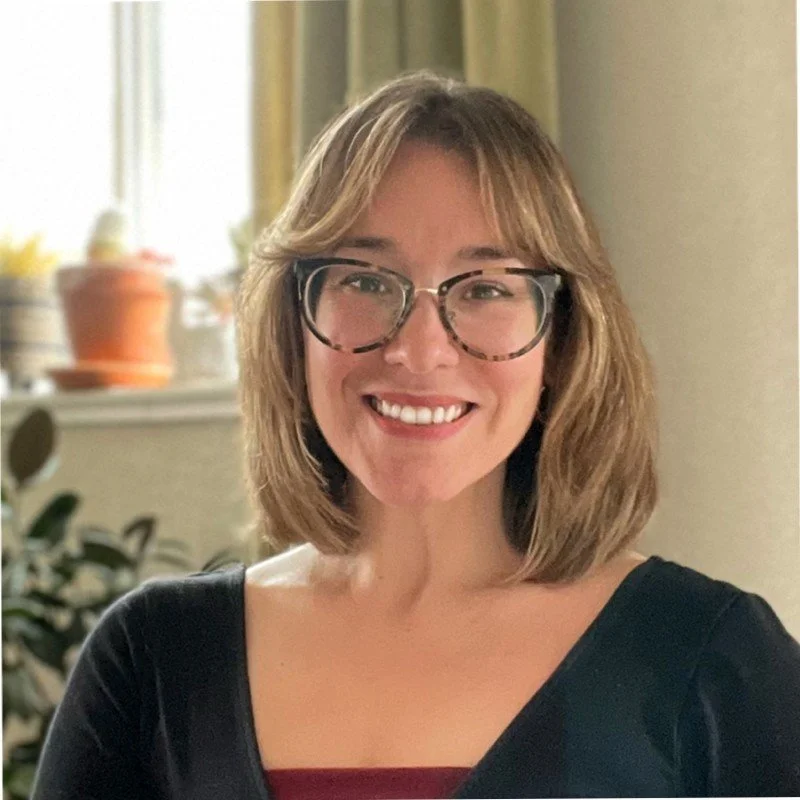 A woman with shoulder-length brown hair, wearing glasses and a black top, smiling indoors with a blurred background of plants and a window.