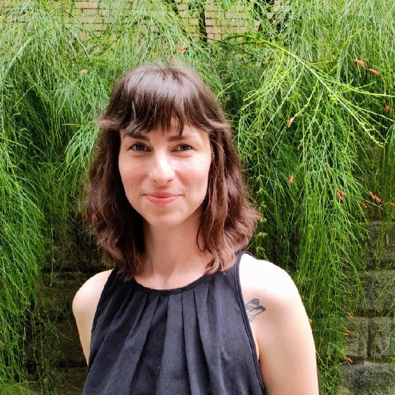 A young woman with shoulder-length brown hair and bangs, wearing a sleeveless black top, standing outdoors in front of lush green foliage.