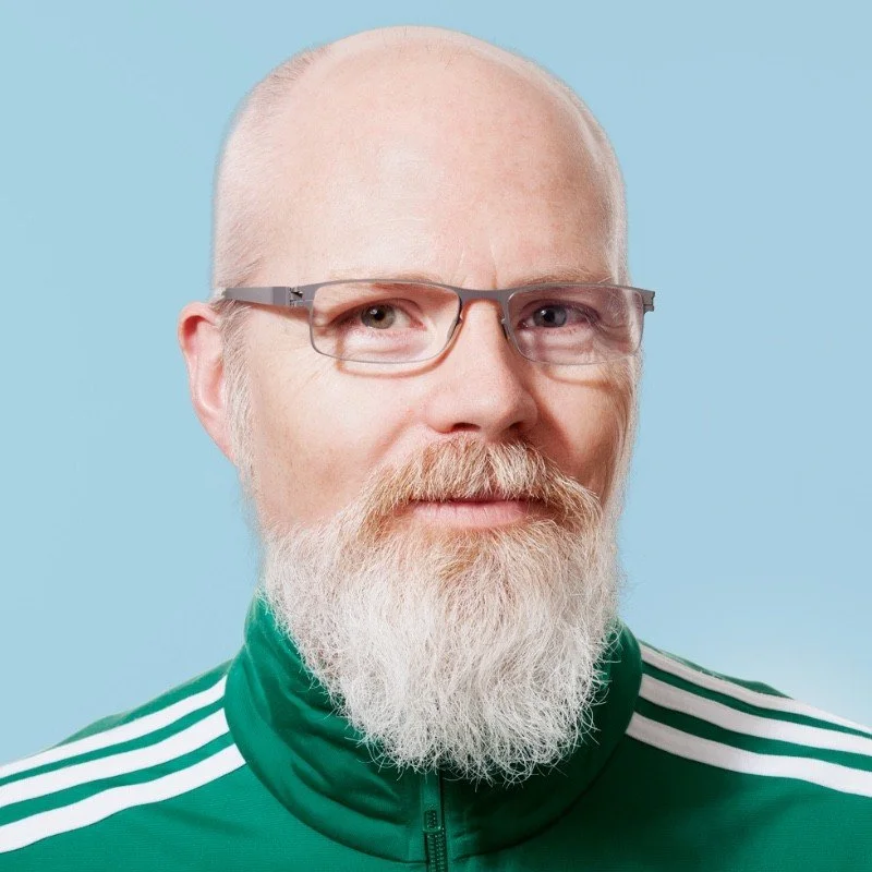 A man with a bald head, glasses, a full white beard, and mustache wearing a green sports jacket with white stripes on the shoulders, against a light blue background.