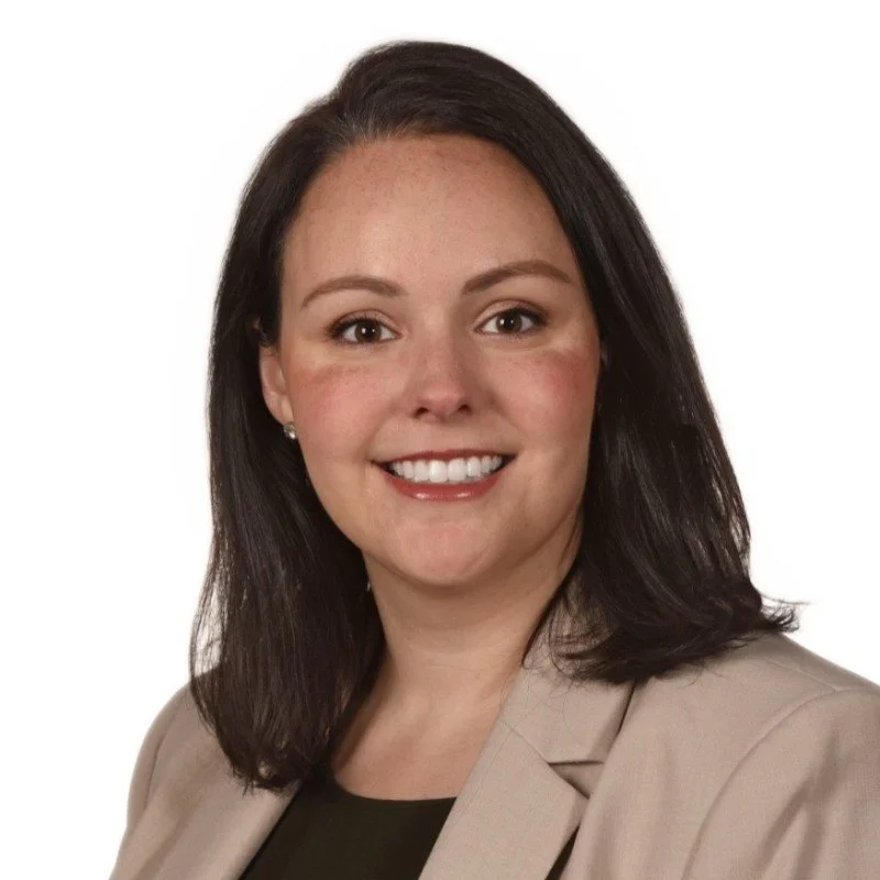 A woman with shoulder-length dark brown hair, smiling, wearing a beige blazer and black top against a white background.