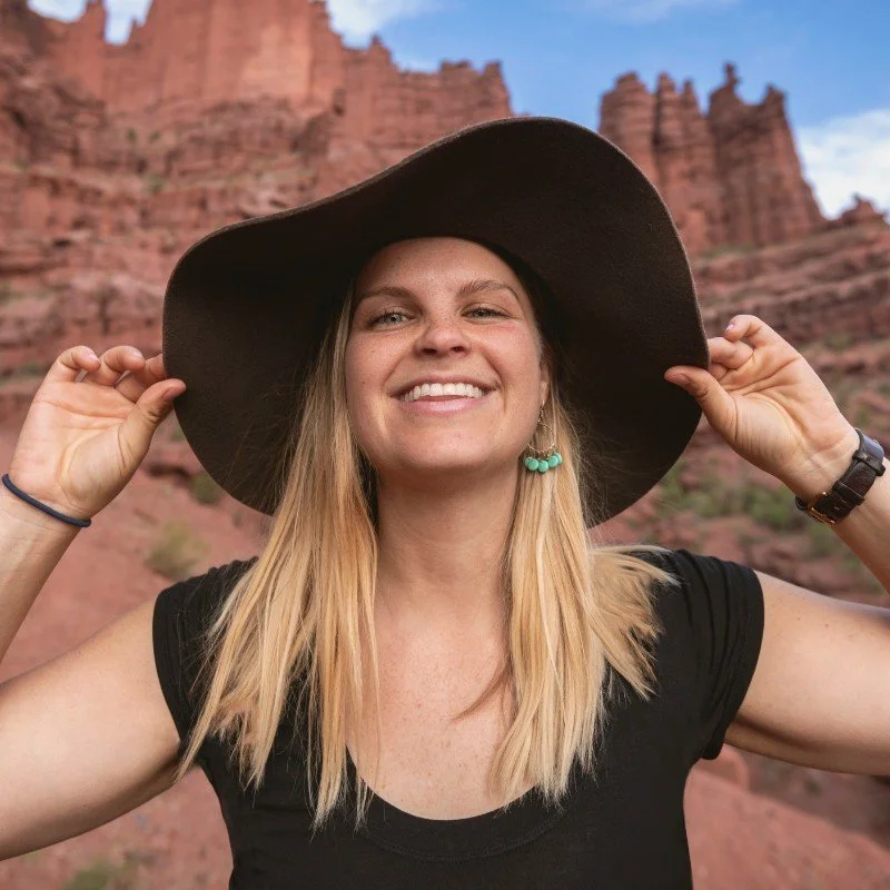 A young woman with blonde hair wearing a large black hat, black t-shirt, and turquoise earrings, smiling in front of red rock formations.