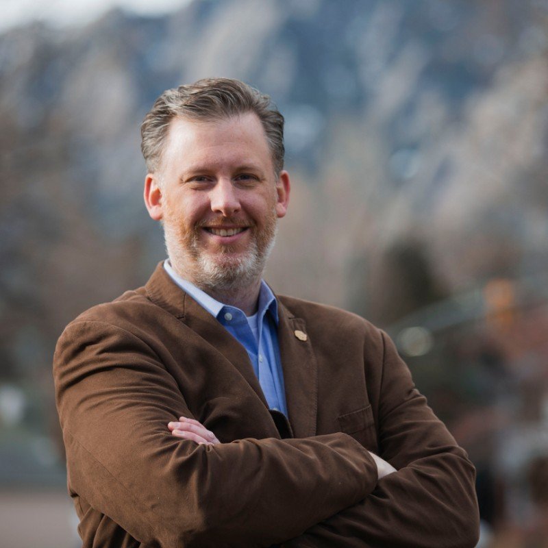 A man with gray hair and beard smiling, standing outdoors with arms crossed, dressed in a blue shirt and brown jacket, with blurred trees in the background.