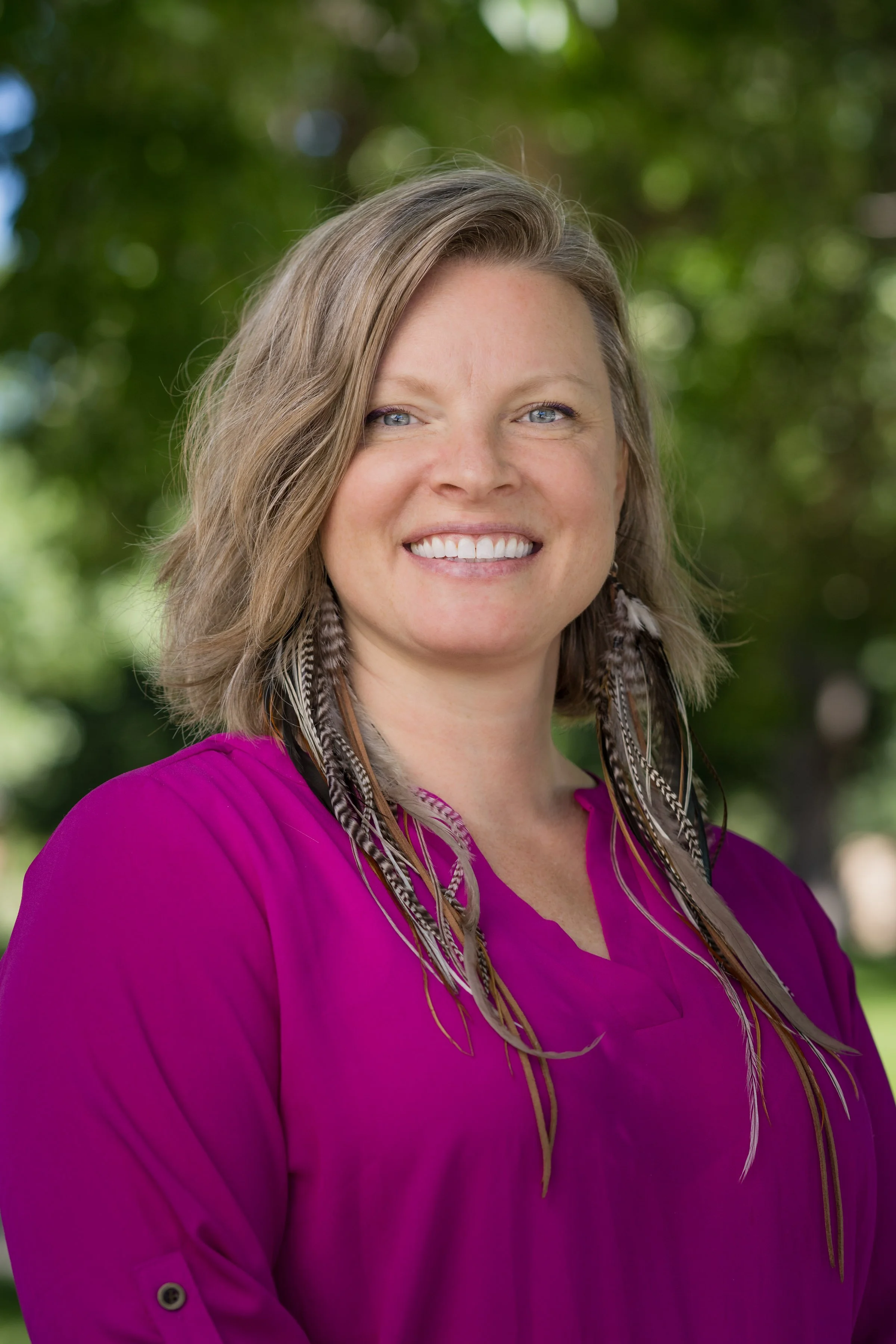 A smiling woman with shoulder-length light brown hair, blue eyes, wearing a magenta blouse and feather earrings, standing outdoors with green trees in the background.