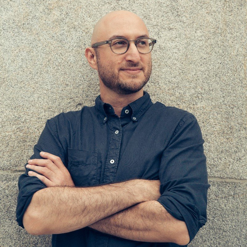 A bald man with glasses and a beard, wearing a dark navy button-up shirt with rolled-up sleeves, standing against a textured stone wall, with arms crossed and looking to the side.