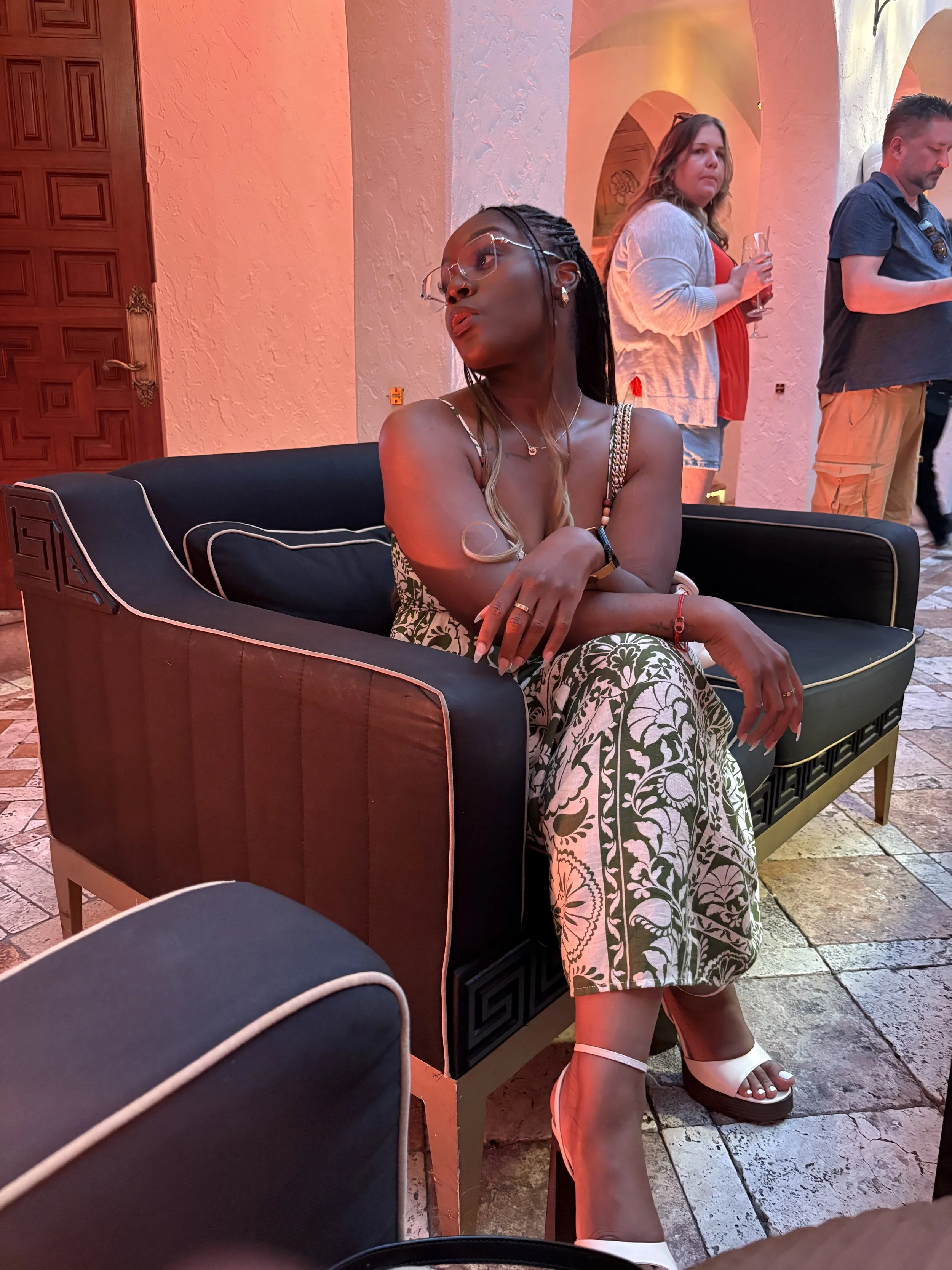 A woman sitting on a black and brown upholstered sofa in a fashionable dress with floral patterns, wearing white high-heeled sandals, at an indoor event with red and warm lighting, with two people standing behind her holding drinks.