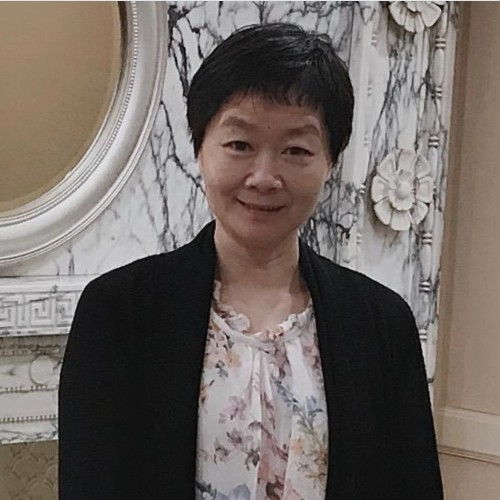 A woman with short black hair smiling, wearing a black jacket over a floral blouse, standing indoors in front of a decorative wall with white flower accents and a mirror.