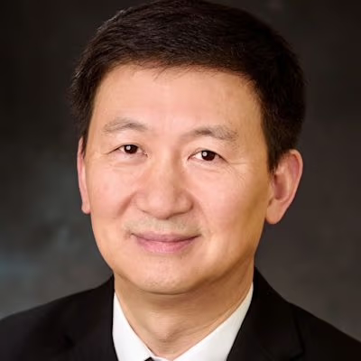 Portrait of a man with short black hair, wearing a black suit and white shirt, smiling against a dark background.