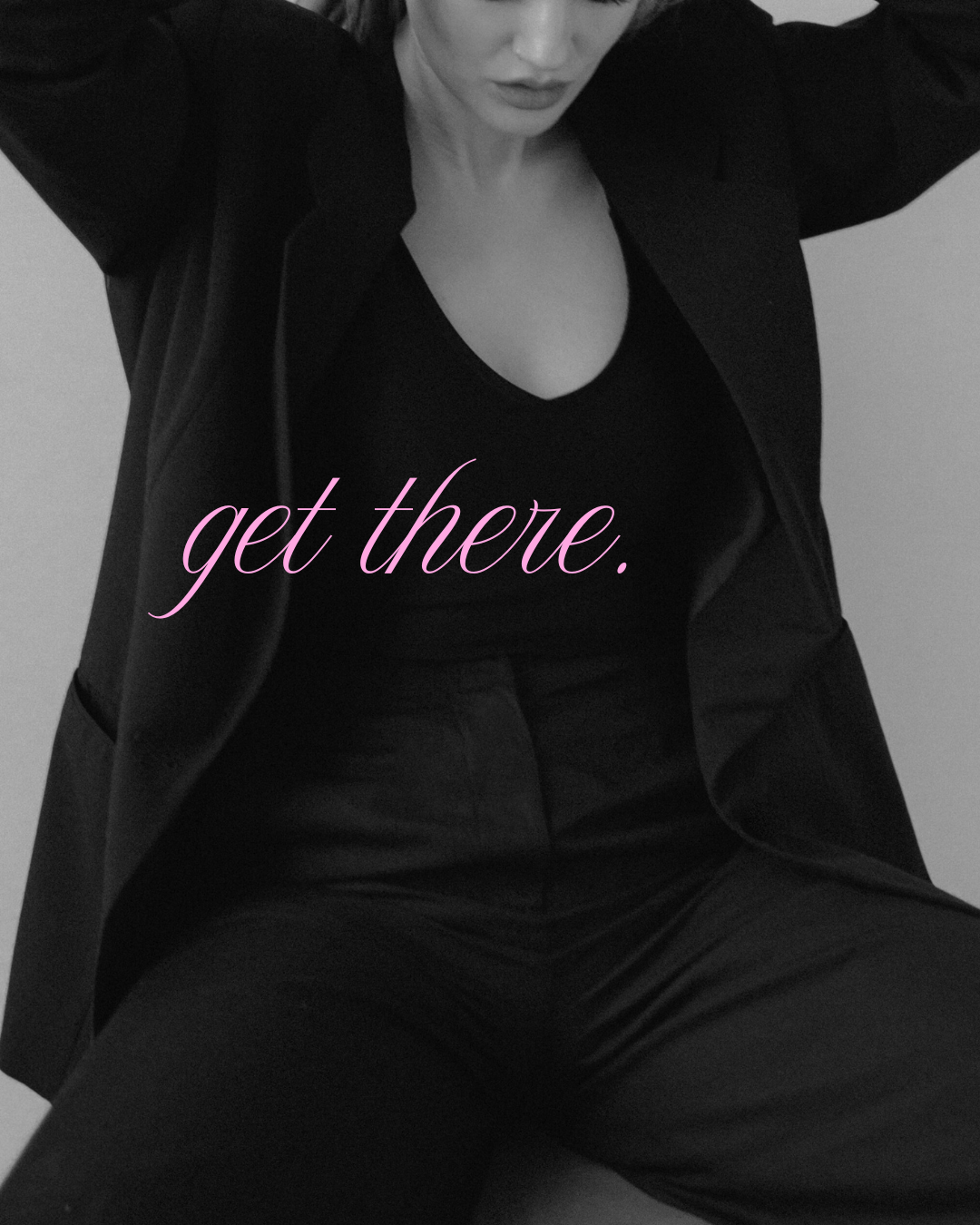 A woman dressed in black clothing with a blazer and pants, sitting with legs apart, with the words "get there." written in pink cursive across her chest.