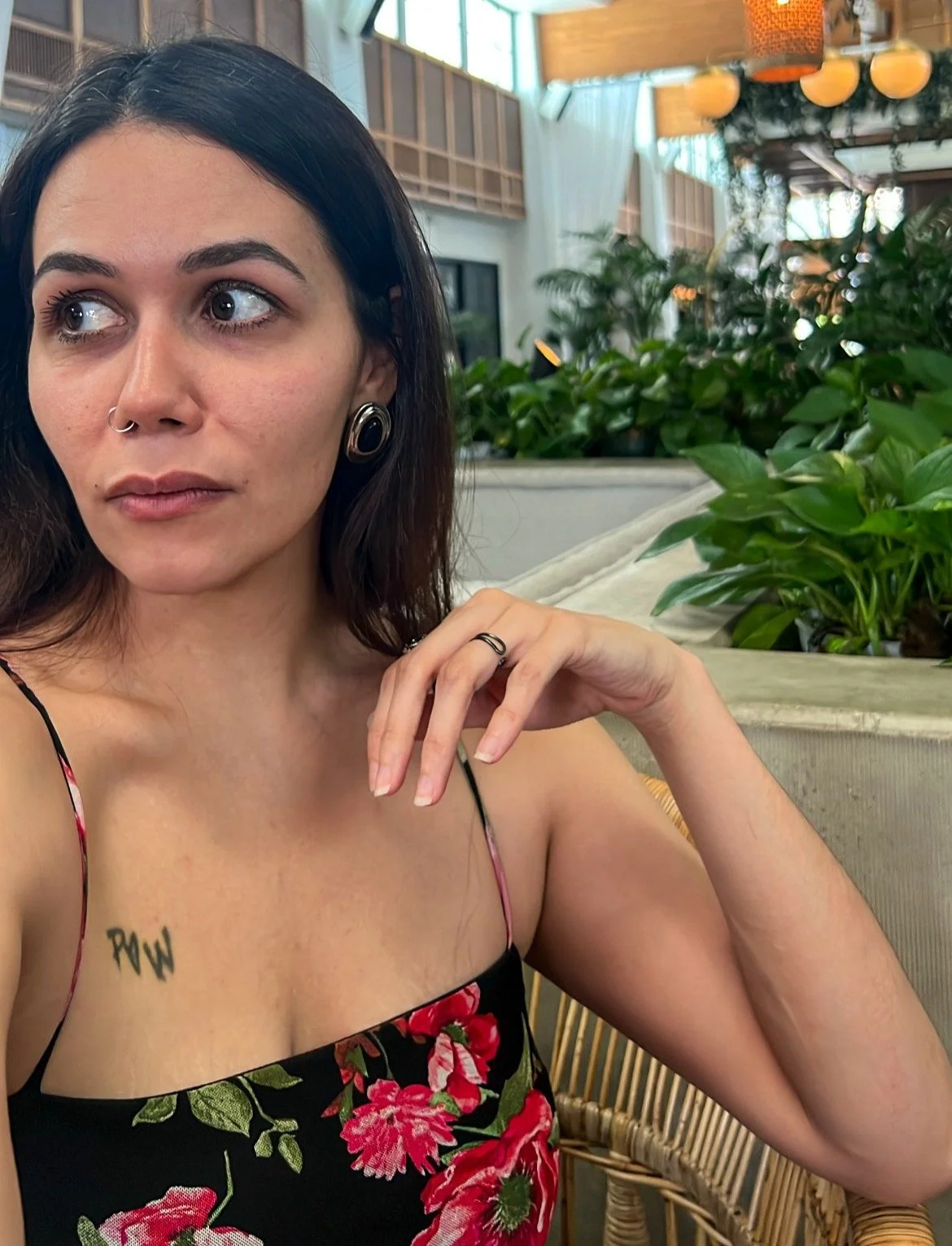 A woman with dark hair and tattoos sitting in a well-lit indoor space with plants and decorative lighting, wearing a floral dress.