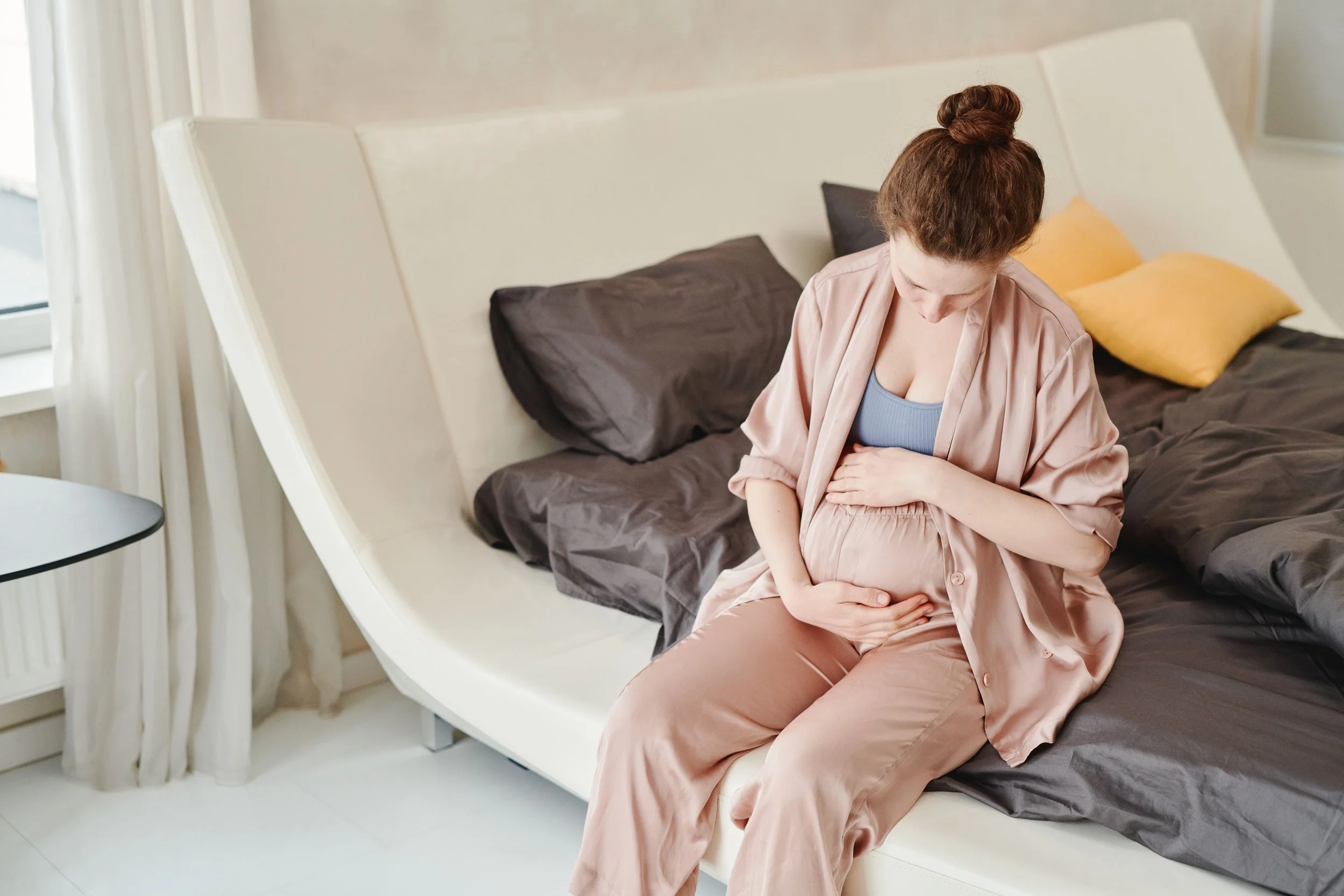 A woman with red hair tied in a bun sitting on a bed, holding her baby bump