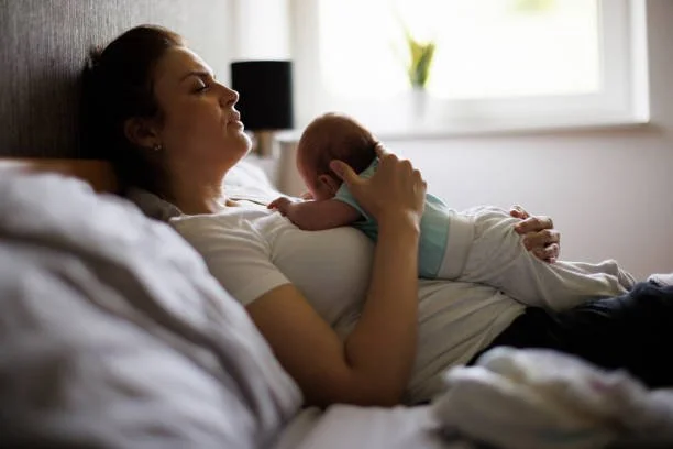 Woman lying on bed with a small child resting on her chest in a bedroom with natural light from a window.