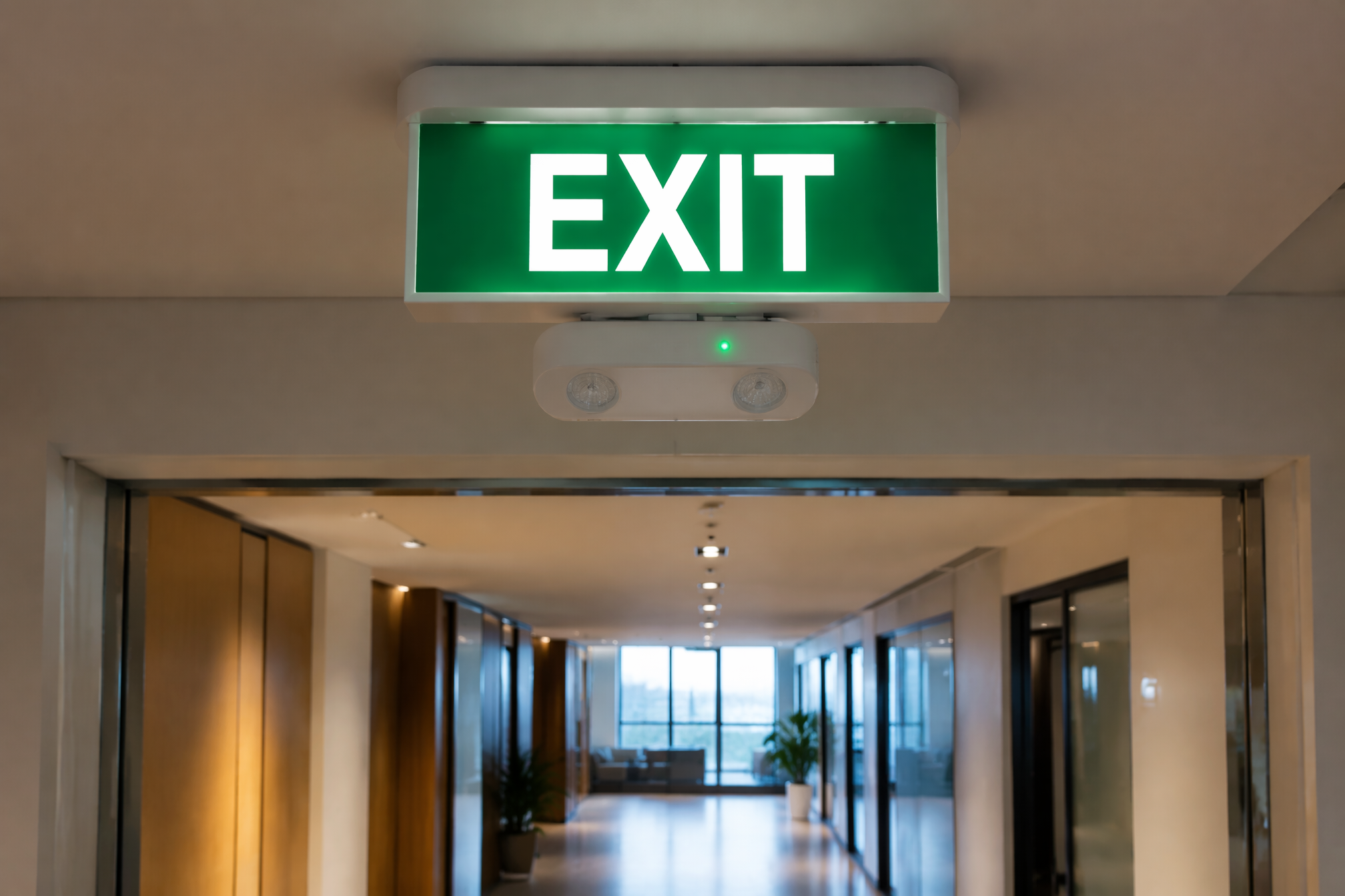 White emergency exit light with green EXIT sign mounted above a corridor doorway in a modern Australian commercial building meeting AS/NZS 2293 compliance