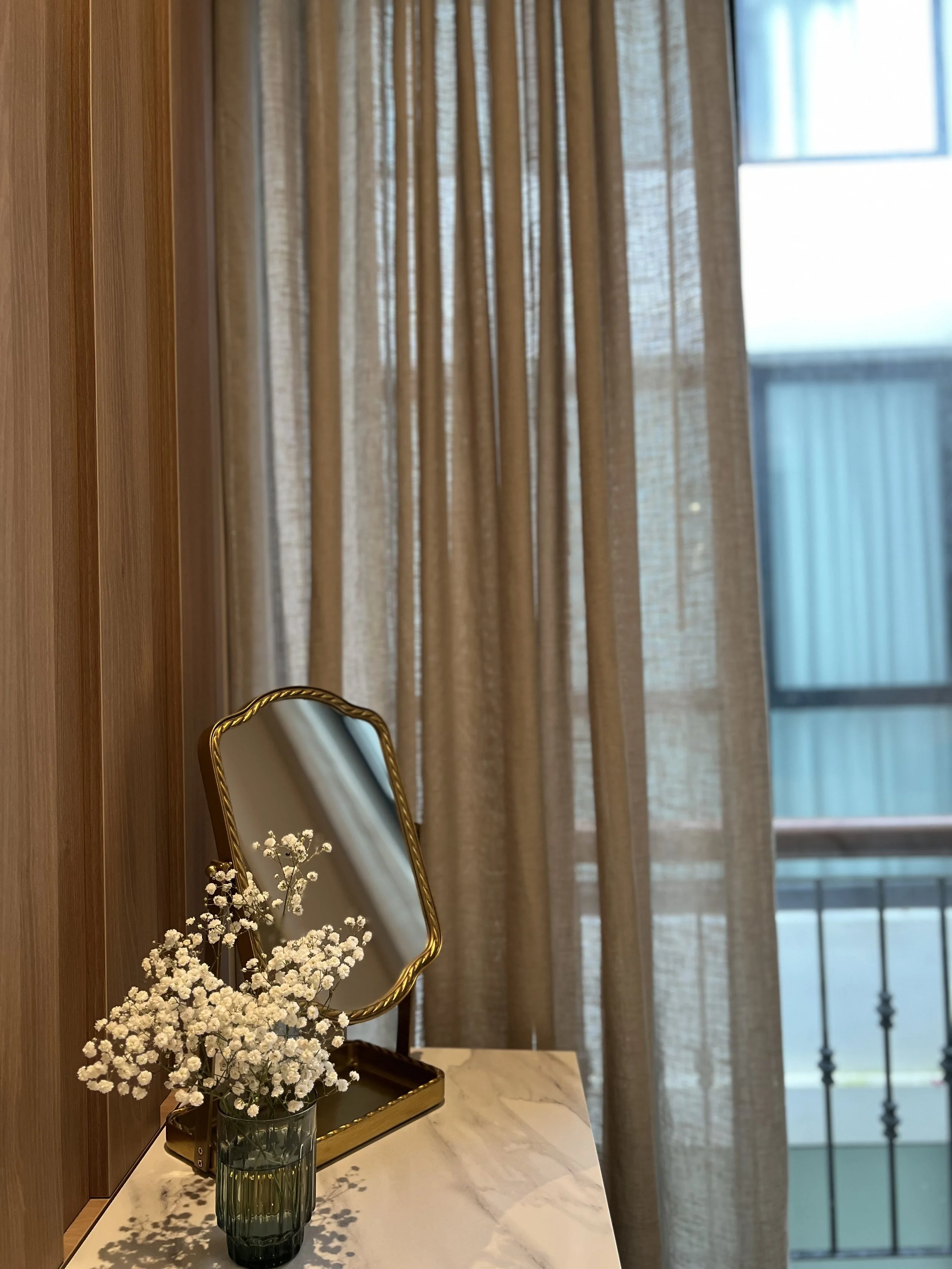 Espejo con marco dorado sobre mesa de mármol, junto a un jarrón con flores blancas, con cortinas beige y ventana con balcón al fondo.