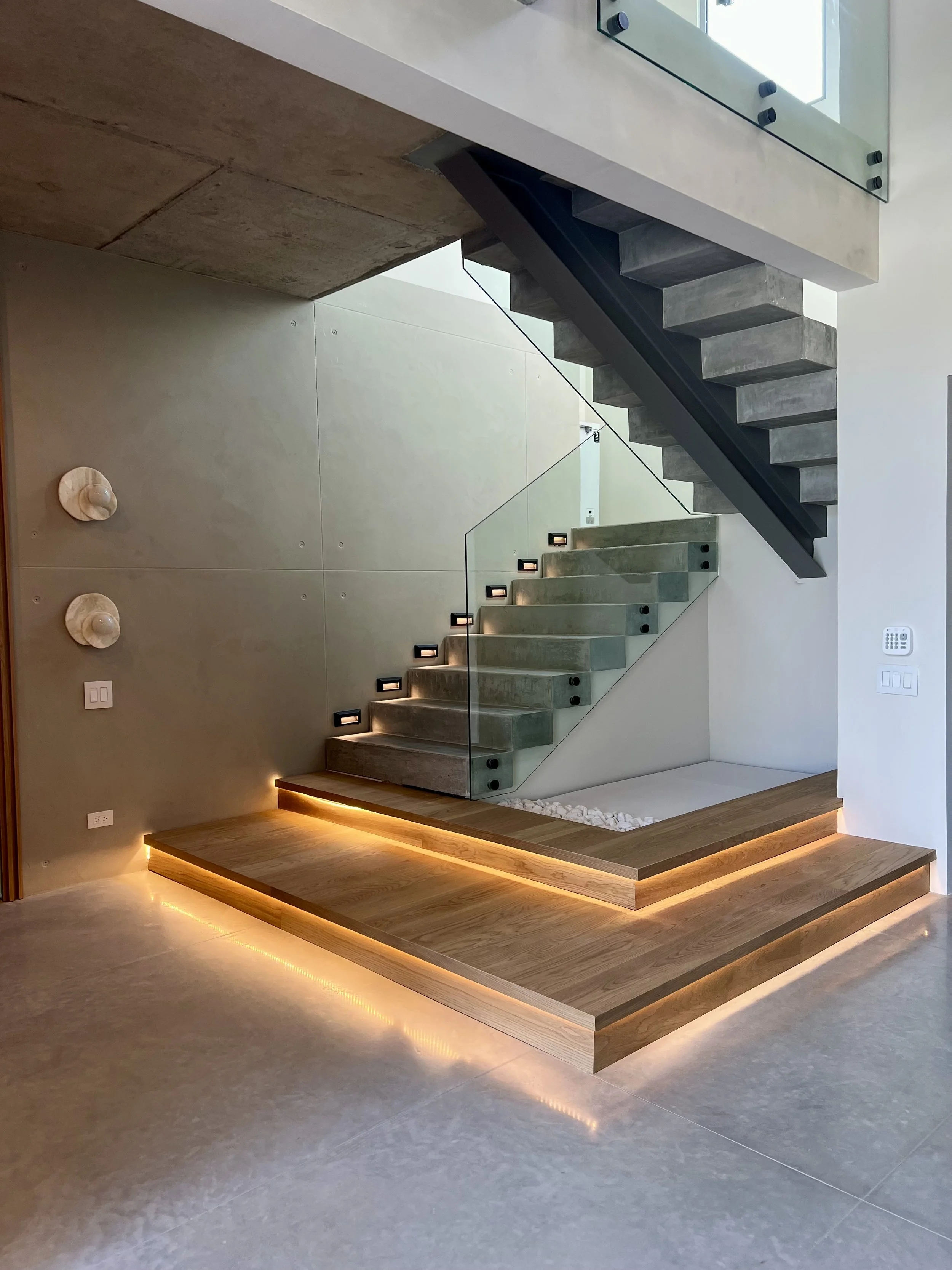 Escaleras modernas de concreto y madera con iluminación LED, barandilla de cristal y pared decorativa en interior de vivienda.