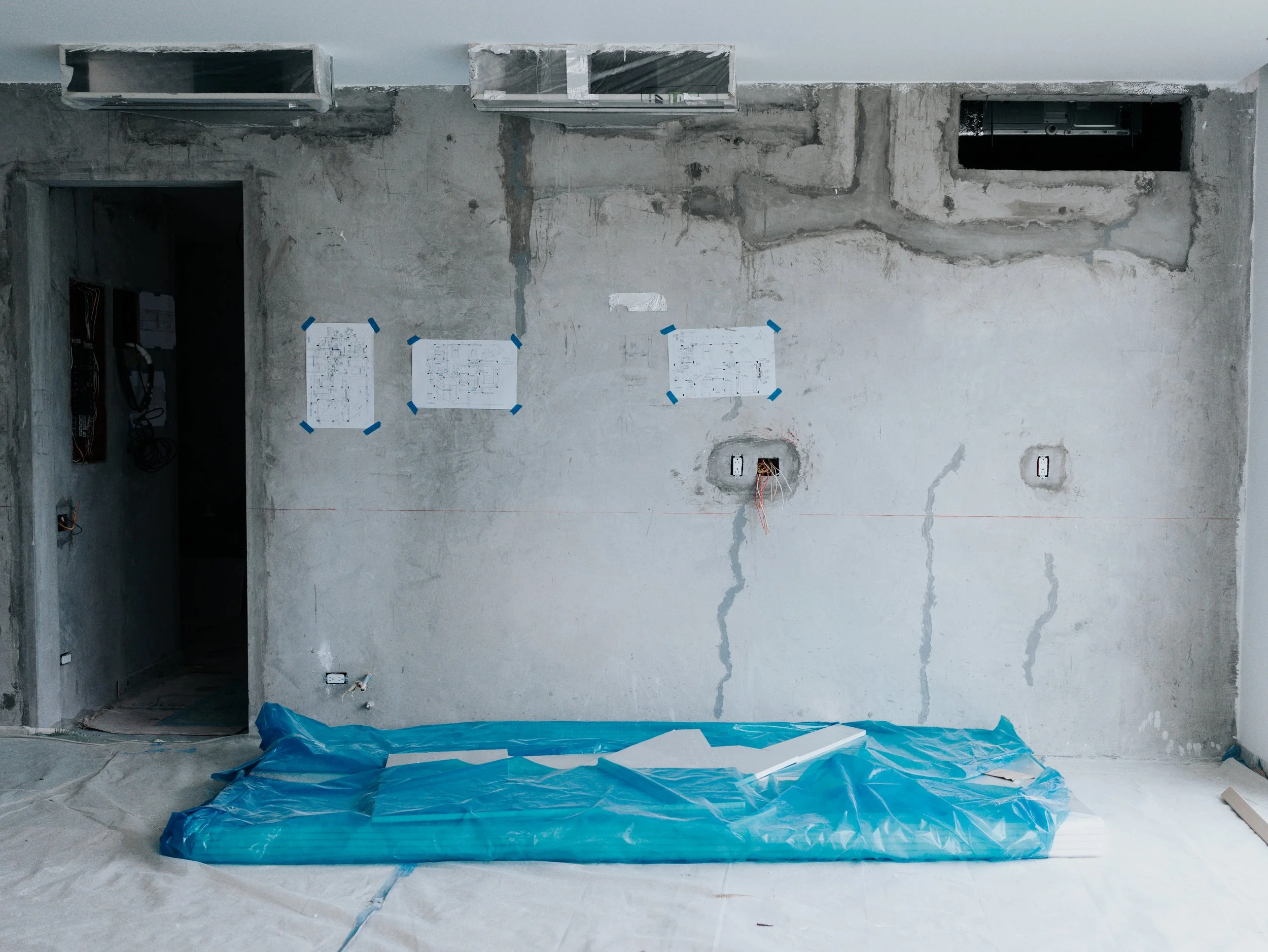 Pared de una construcción en proceso con cables expuestos y planos en la pared, cubierta con plástico azul y papel, en un ambiente de obra