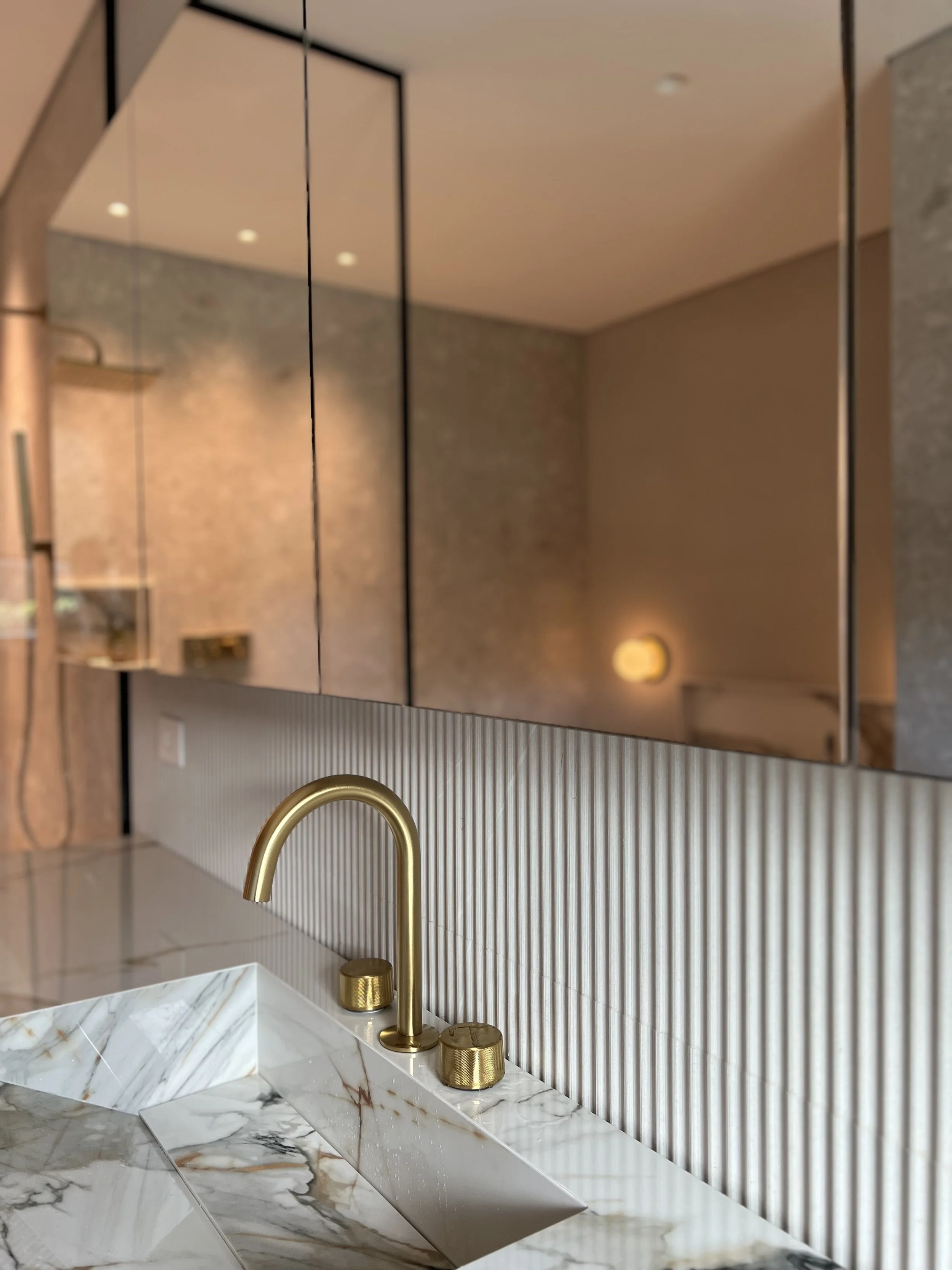 Imagen de una cocina moderna con mesón de mármol blanco con vetas doradas, grifo de oro y espejos que reflejan la pared decorada con mosaico, luces cálidas en el techo y una pared con papel texturizado