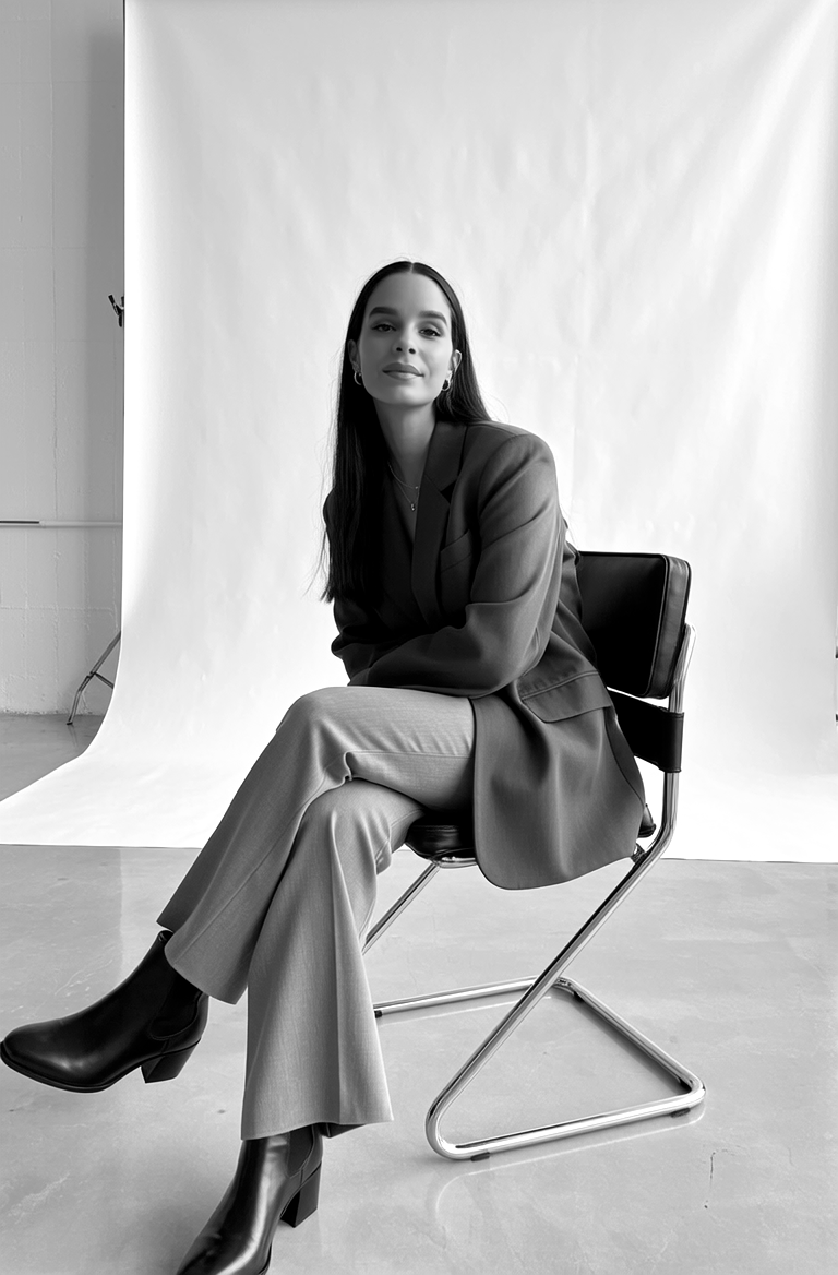 Mujer sentada en una silla de estudio, usando traje sastre y botas negras, en un fondo de estudio fotográfico en blanco y negro.