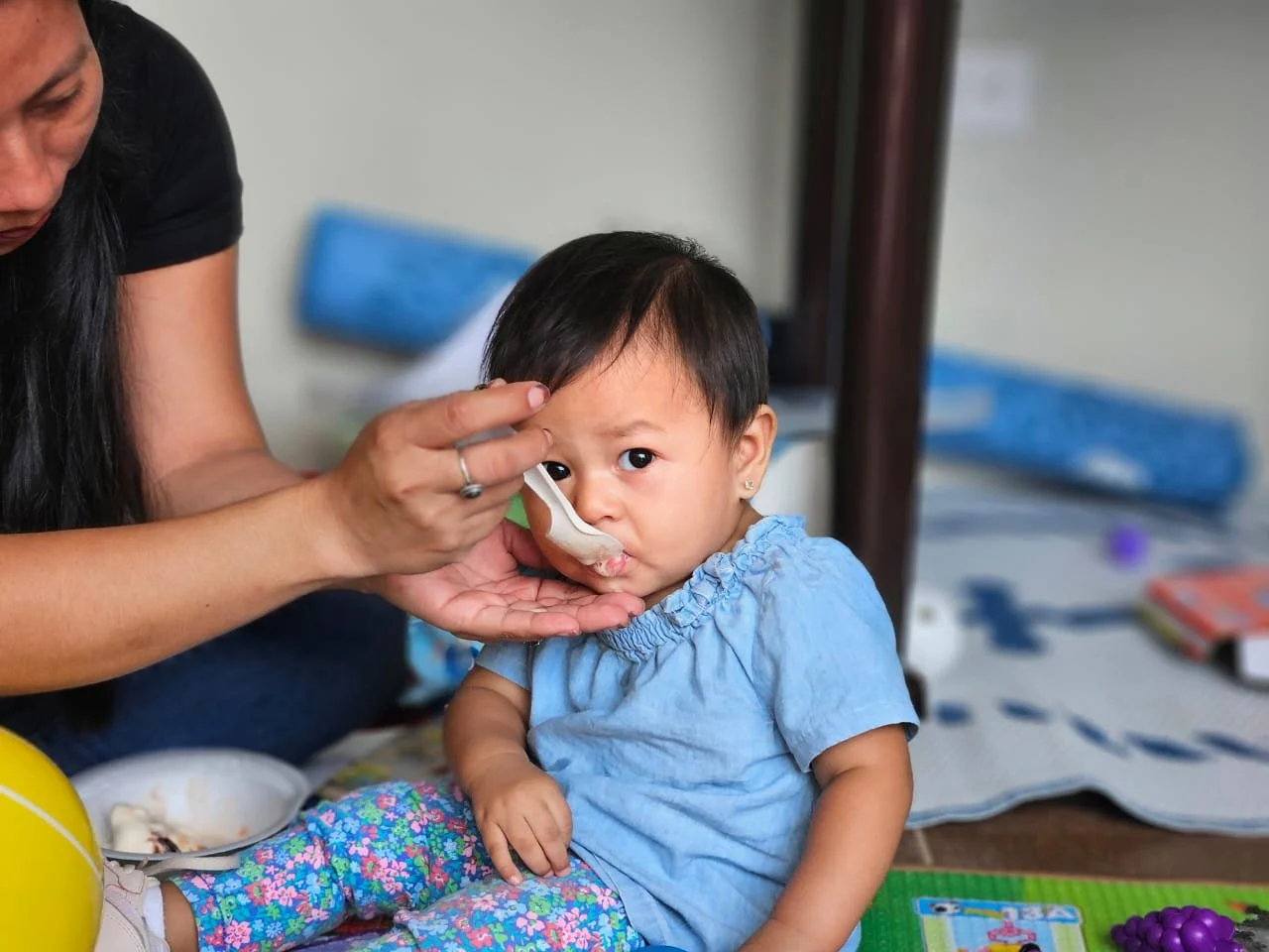 A young girl with a spoon being fed by an adult woman, sitting on the floor with toys and books around.